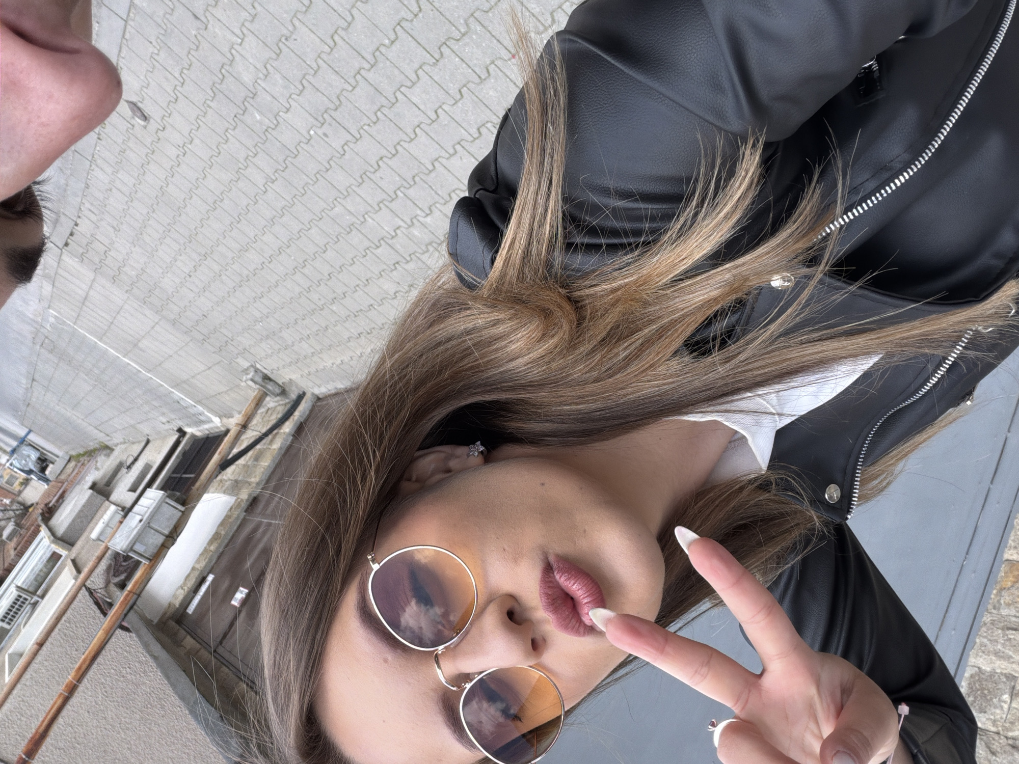 A photo of a woman with long brown hair wearing a black leather jacket, a white shirt, and round sunglasses in a city environment, likely Panagyurishte, Bulgaria. She is making a peace sign with her right hand. The background features buildings with weathered facades and a tiled pedestrian walkway, suggesting an urban setting in Bulgaria. The sunglasses reflect the sky and clouds.