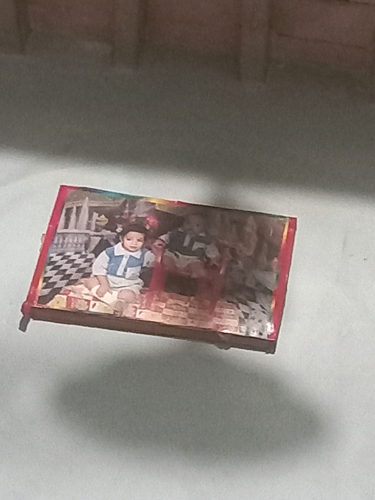 The image shows a framed photograph displayed on a light-colored, textured surface. The photograph features a young child, likely a boy, sitting on the floor. He is wearing a blue and white collared shirt and white pants. Behind him, there appears to be a black and white checkered pattern on the floor and some architectural elements that suggest an indoor setting. Another figure, also a child, is partially visible in the background, seated in a chair. The photograph is framed with a red border. The overall lighting is dim, casting a shadow of the framed photograph onto the surface below. The context provided places this scene in the city of Jalandhar, India.