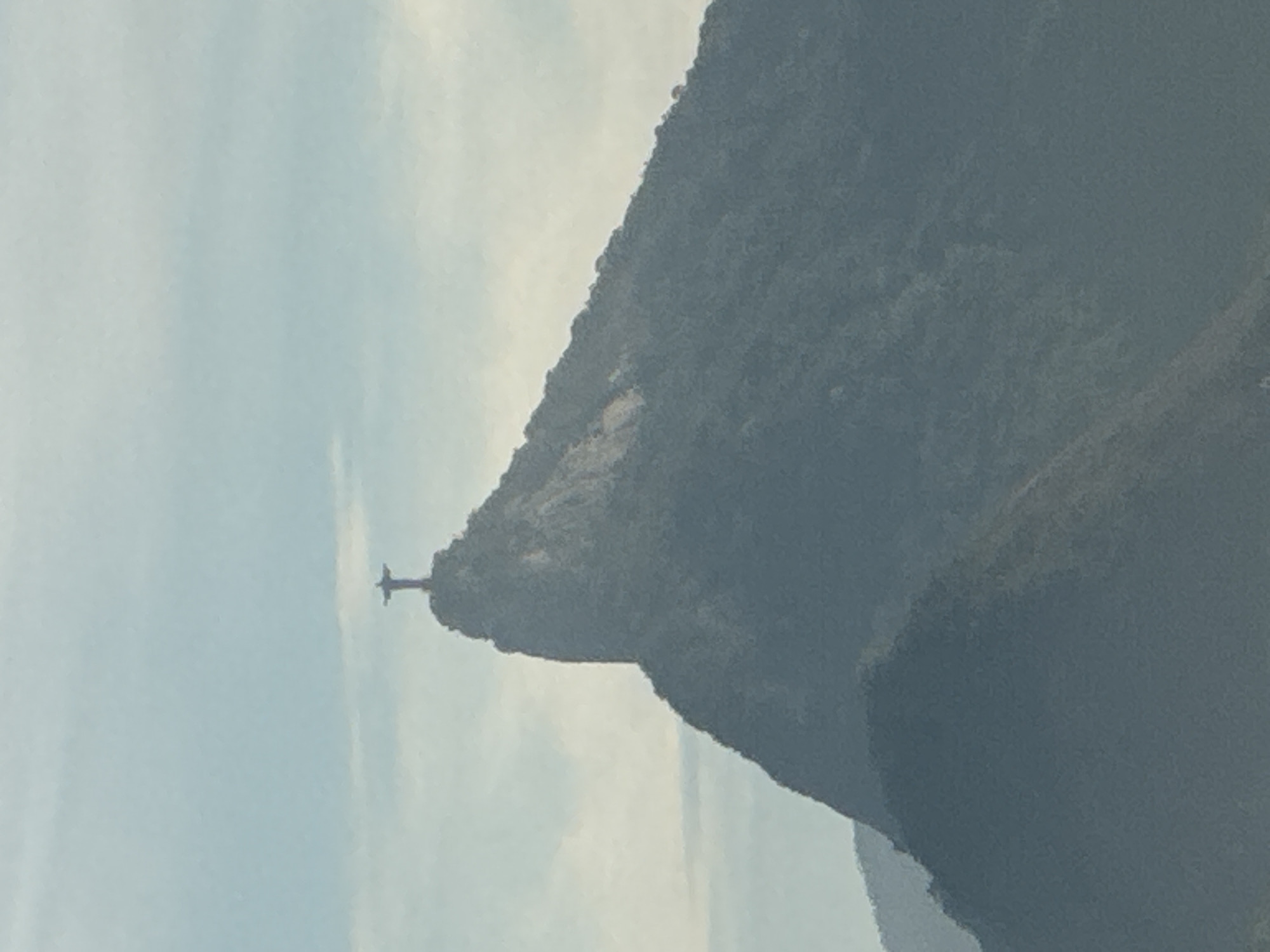 The image shows a silhouette of the Christ the Redeemer statue atop Corcovado Mountain in Rio de Janeiro, Brazil. The statue, a colossal Art Deco statue of Jesus Christ, is a global icon of Christianity and a cultural icon of both Rio de Janeiro and Brazil. It is situated at the peak of the Corcovado mountain, overlooking the city. The mountain itself is steep and rocky, covered in dense vegetation. The sky above is a pale blue with scattered white clouds. The overall impression is one of awe and grandeur, with the statue standing sentinel over the city.