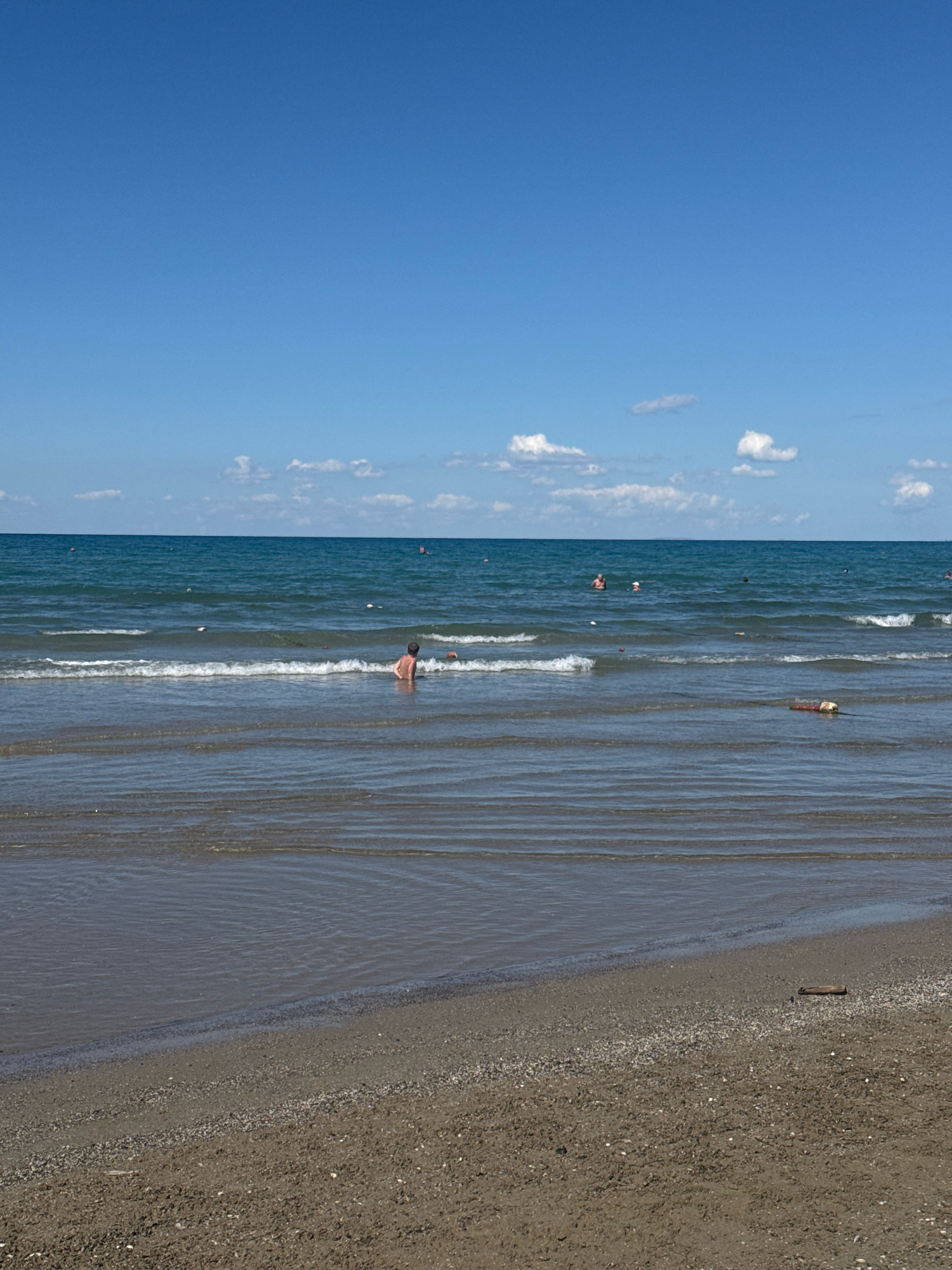 The image depicts a beach scene at the city of Anapa, Russia. A clear blue sky with a few scattered white clouds stretches above a calm blue sea. Gentle waves lap the sandy shore. In the foreground, the wet sand shows a pattern of receding water. Several individuals are visible in the water, some appearing to be swimming or wading. One person, a child, is seated in the shallow water closer to the shore. Further out in the sea, there are some buoys or floating markers. A piece of driftwood lies on the sand near the water's edge. The overall atmosphere is serene and suggests a pleasant day at the beach.