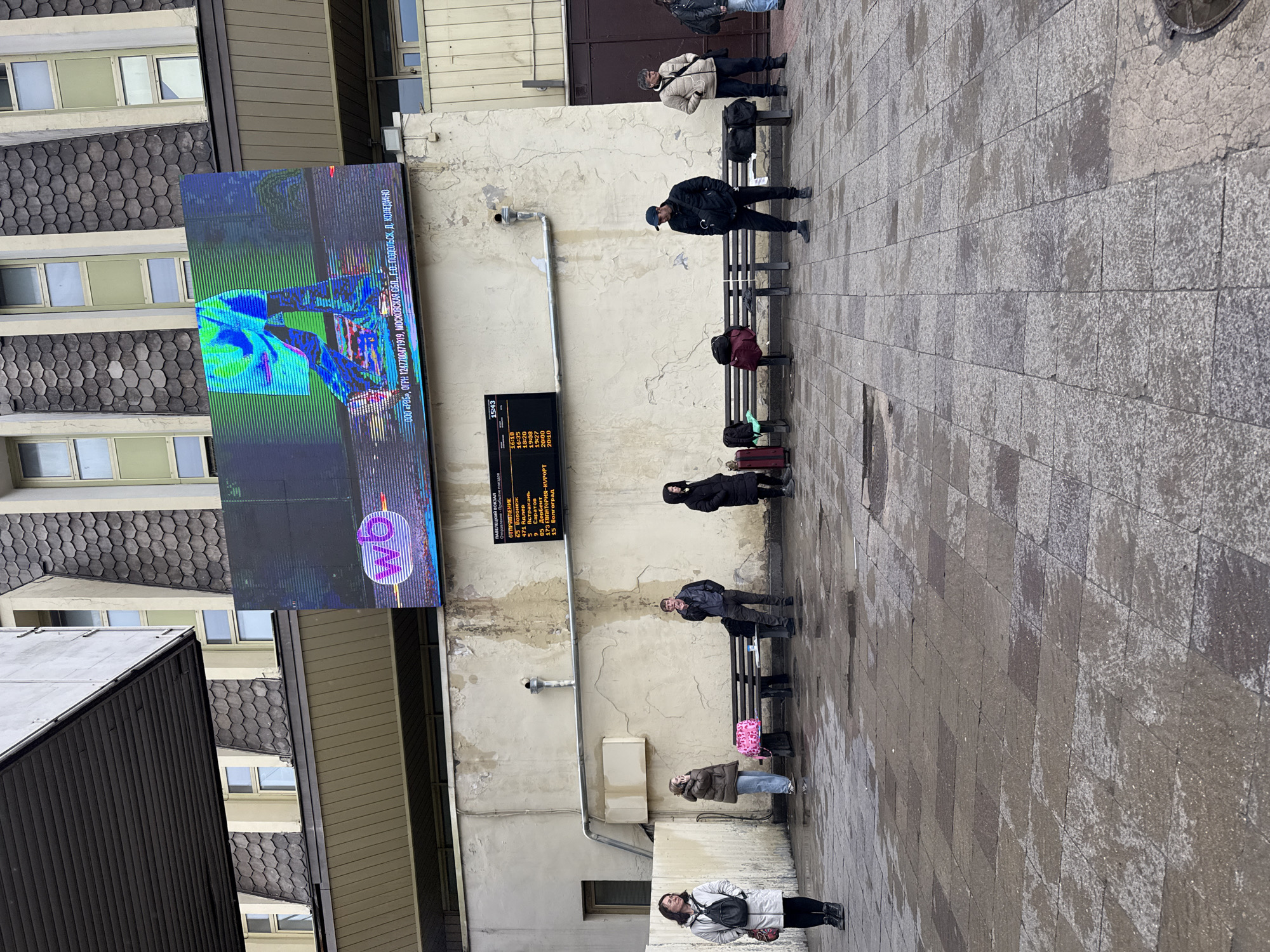 The image depicts an outdoor scene in what appears to be a train station or waiting area. Several people are either sitting on benches or standing along a wall, likely waiting for transportation. There's a large digital screen displaying information, possibly related to train schedules or advertisements. The screen shows a distorted image and the logo for "wb". A smaller display shows arrival and departure times for various destinations, including Voronezh, Adler, Astrakhan, Saratov, and others. The building behind the waiting area has a mix of architectural styles, with windows and different wall textures. The ground is paved with rectangular tiles. The overall atmosphere suggests a public transportation hub in a somewhat urban environment. The OCR provides additional details, including a company name and registration number.