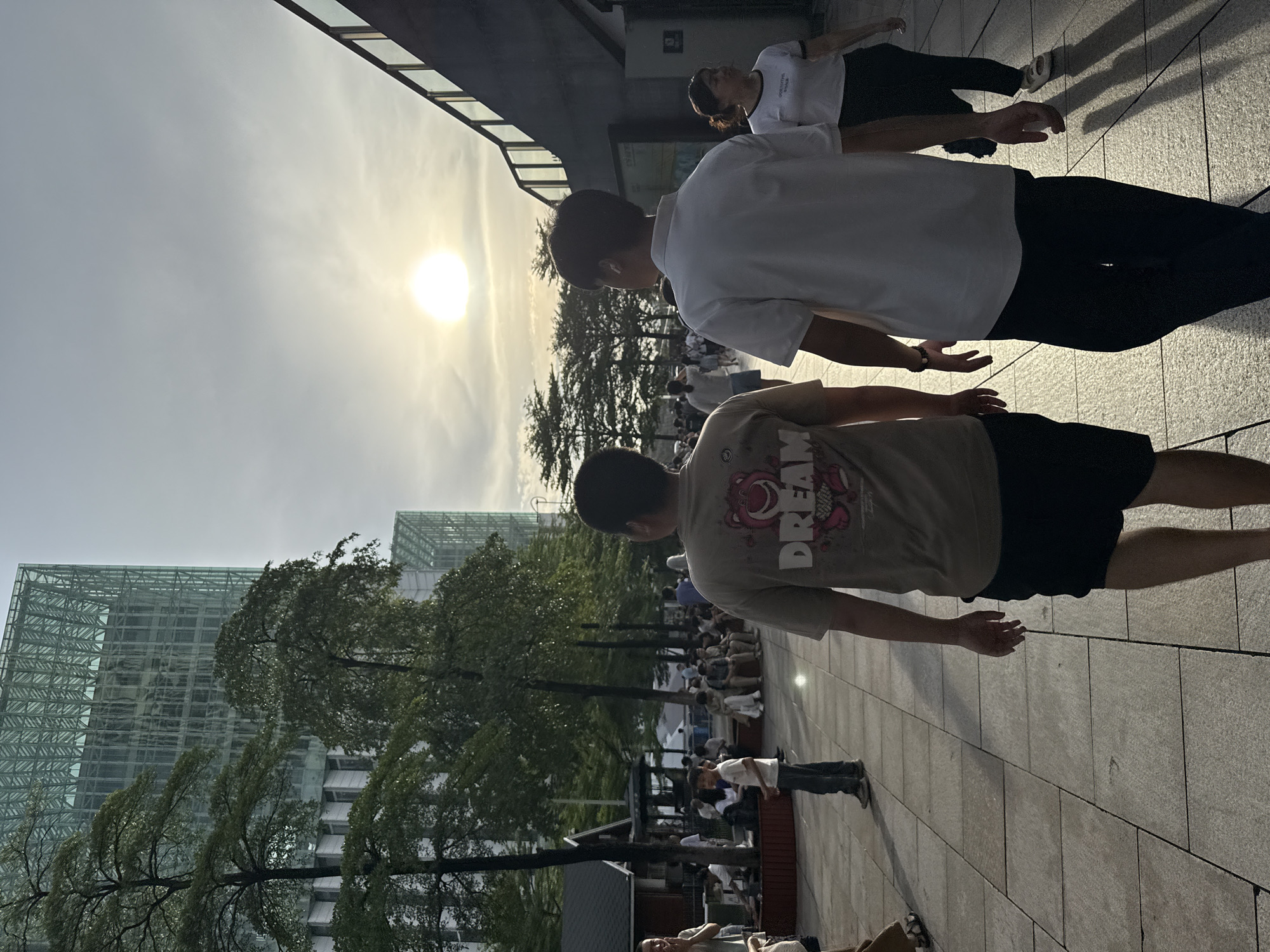 The image captures a scene in the Haizhu District, China, likely during late afternoon or early evening, given the low angle of the sun casting long shadows. In the foreground, two individuals are walking away from the camera. The person on the left wears a tan t-shirt with a graphic design featuring a cartoon character and the word "DREAM," and black shorts. The person on the right wears a white polo shirt and dark pants. Behind them, other people are visible walking and sitting, suggesting a public outdoor space, possibly a plaza or park area adjacent to commercial buildings.
In the background, a modern, multi-story building with a glass facade and a distinctive green structural grid rises prominently. Trees with lush green foliage are interspersed between the buildings and the pedestrian area. To the right of the frame, a dark, elevated structure, possibly a walkway or part of a building, features a railing with glass panels. The sky is bright with the sun visible, partially obscured by thin clouds, indicating a clear or partly cloudy sky. The ground is paved with rectangular tiles. The overall atmosphere is one of urban activity and leisure.