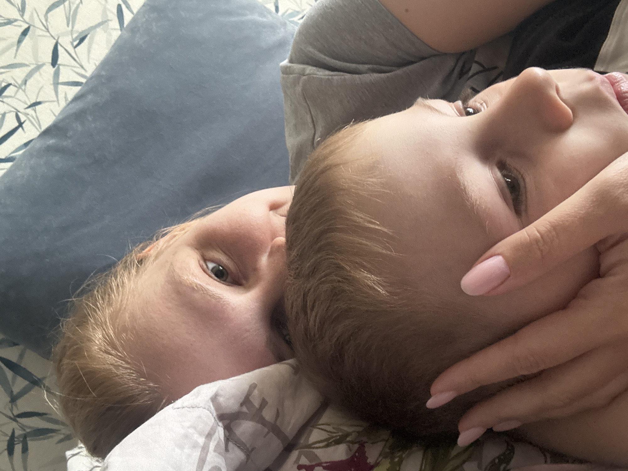 A close-up, angled shot depicts a woman and child lying on a bed. The woman, with light skin and light brown hair, looks directly at the camera. The child, also light-skinned with light brown hair, lies close to the woman. A hand with manicured fingernails rests on the child's face. They are lying on a bed with a patterned sheet and a blue pillow behind the woman. Location information is not discernible from the image.