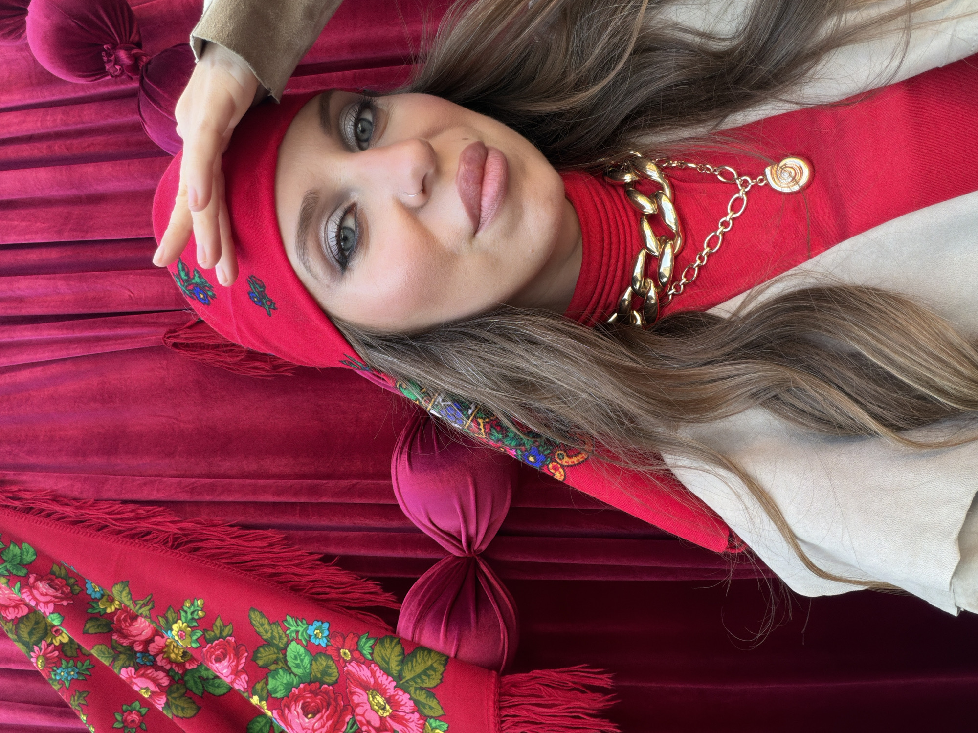 A high-angle portrait features a young woman lying down, gazing directly at the viewer with a poised and confident expression. Her long, wavy brown hair cascades around her head, framing her fair skin, striking blue eyes, and light pink lips. She wears a small nose ring and noticeable makeup, including eyeshadow and eyeliner.
She is dressed in a vibrant red headscarf, which is intricately embroidered with green leaves, pink roses, and blue and yellow floral motifs. A thick, gold-colored chain necklace with a circular pendant rests against her red high-necked top. A light beige or cream-colored outer garment is visible on her right shoulder. Her left hand is gently placed on her headscarf near her temple.
The background is rich with various red textiles. A deep red, possibly velvet, fabric is draped with soft folds, covering much of the setting. A larger, fringed red textile, adorned with a floral pattern matching her headscarf, is visible in the lower left. Two bunched or tied fabric elements, also in a deep red, appear in the upper left background, contributing to the textured and stylized indoor environment. No text is visible in the image.
