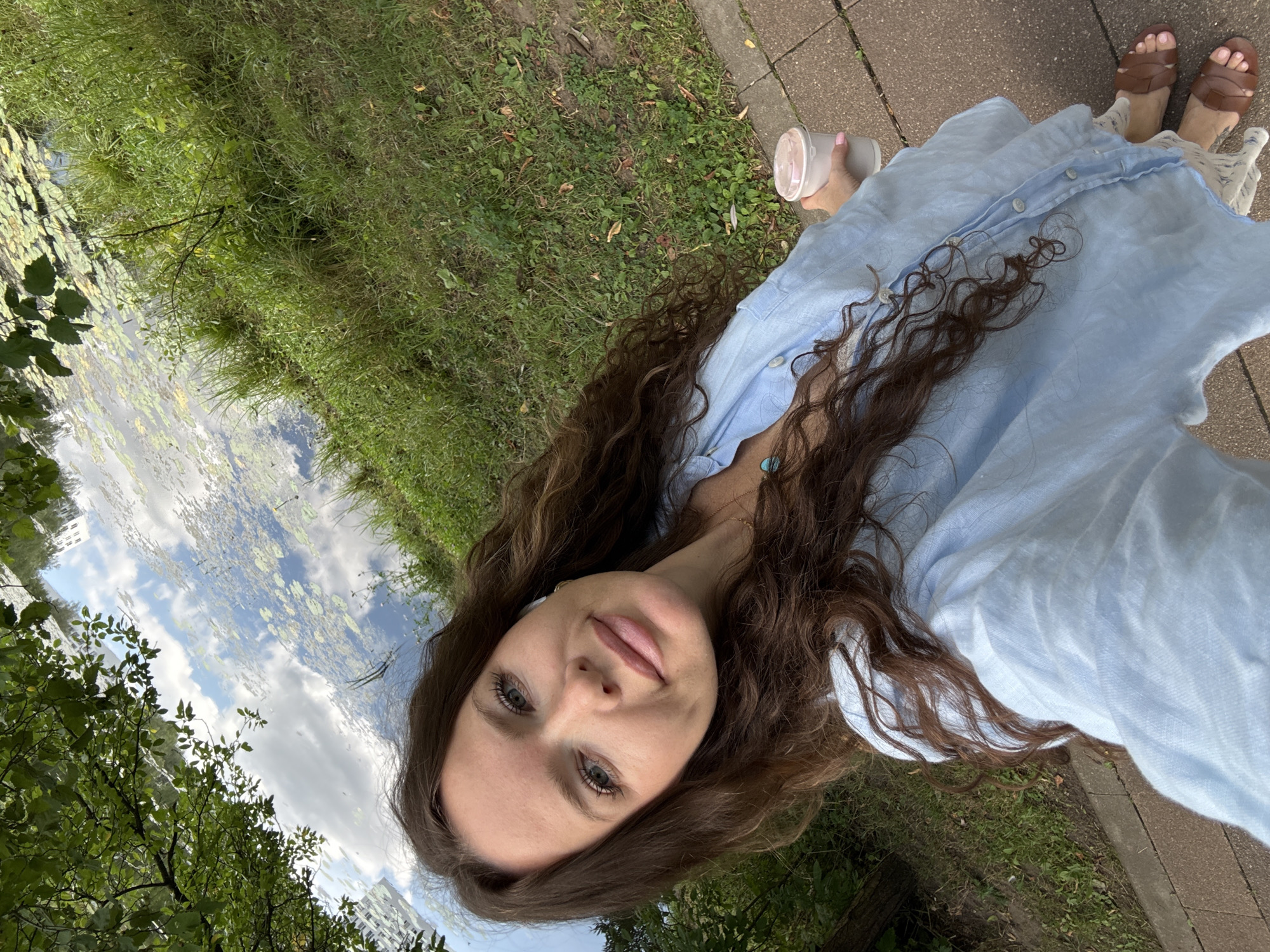 A young woman with long, curly brown hair and fair skin is taking a selfie outdoors. She is wearing a light blue, button-down shirt and white patterned shorts, with brown sandals on her feet. A turquoise pendant necklace is visible. She is holding a light pink disposable cup with a lid in her right hand. The background shows a body of water with lily pads, reflected sky and clouds, lush green grass, and trees. The scene appears to be a park or waterfront area, possibly in Moscow, Russia. The lighting suggests it is daytime.