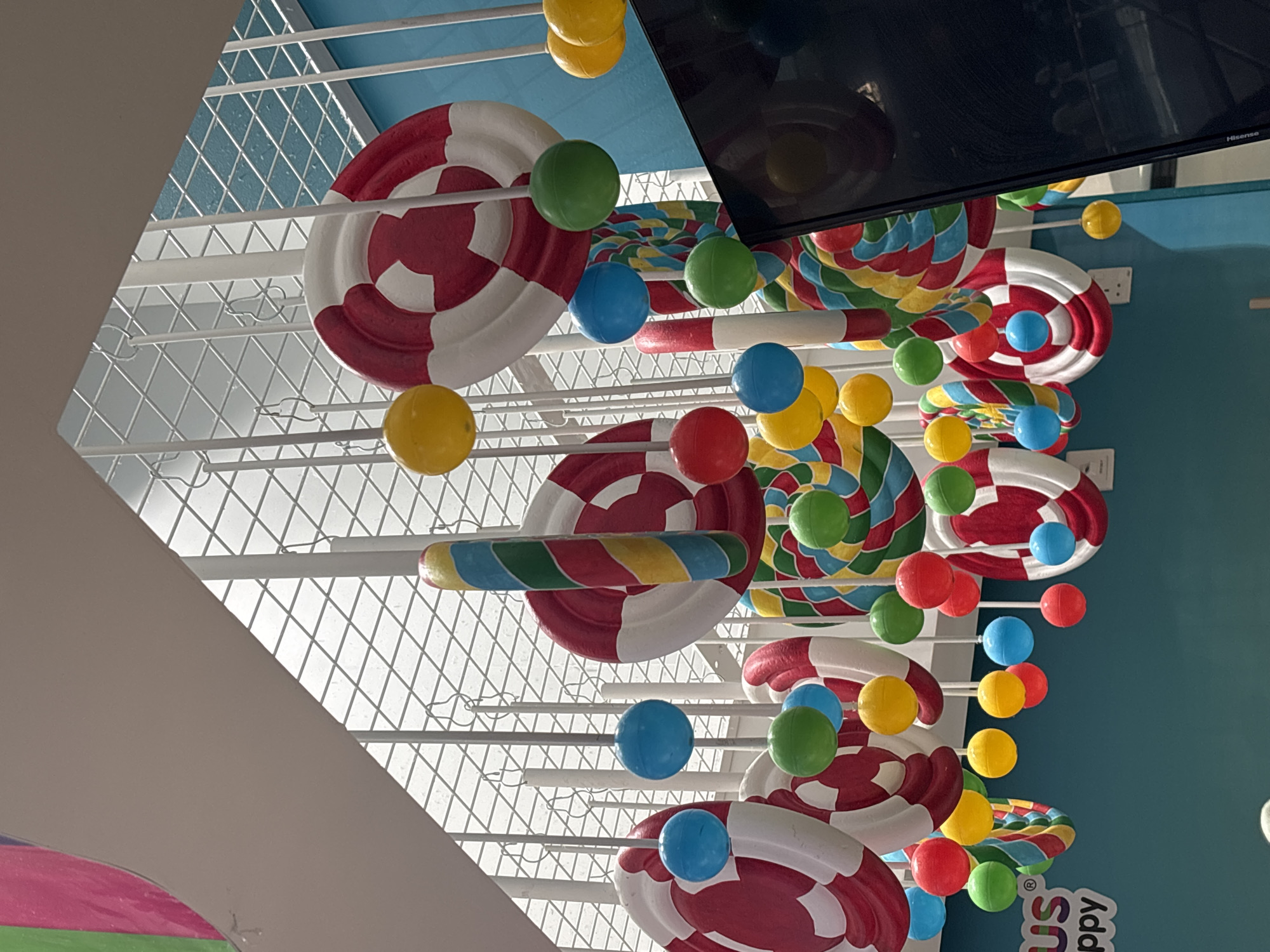 This image shows a colorful display of oversized candy decorations suspended from the ceiling in a brightly lit indoor space. The decorations include large, circular lollipops with red and white stripes, and vertically striped candy canes. Smaller, spherical ornaments in various colors such as blue, green, yellow, and red are also hanging at different levels. The background features a white grid ceiling structure and a teal-colored wall. A portion of a black screen, possibly a television or monitor, is visible on the right side of the frame. Text that reads "US" and "ppy" is partially visible in the lower left corner, suggesting this might be a children's entertainment venue or themed area. The overall impression is one of a playful and festive environment. While the image does not explicitly state its location, such a decorative style is common in family-friendly entertainment centers found in Dubai, United Arab Emirates.