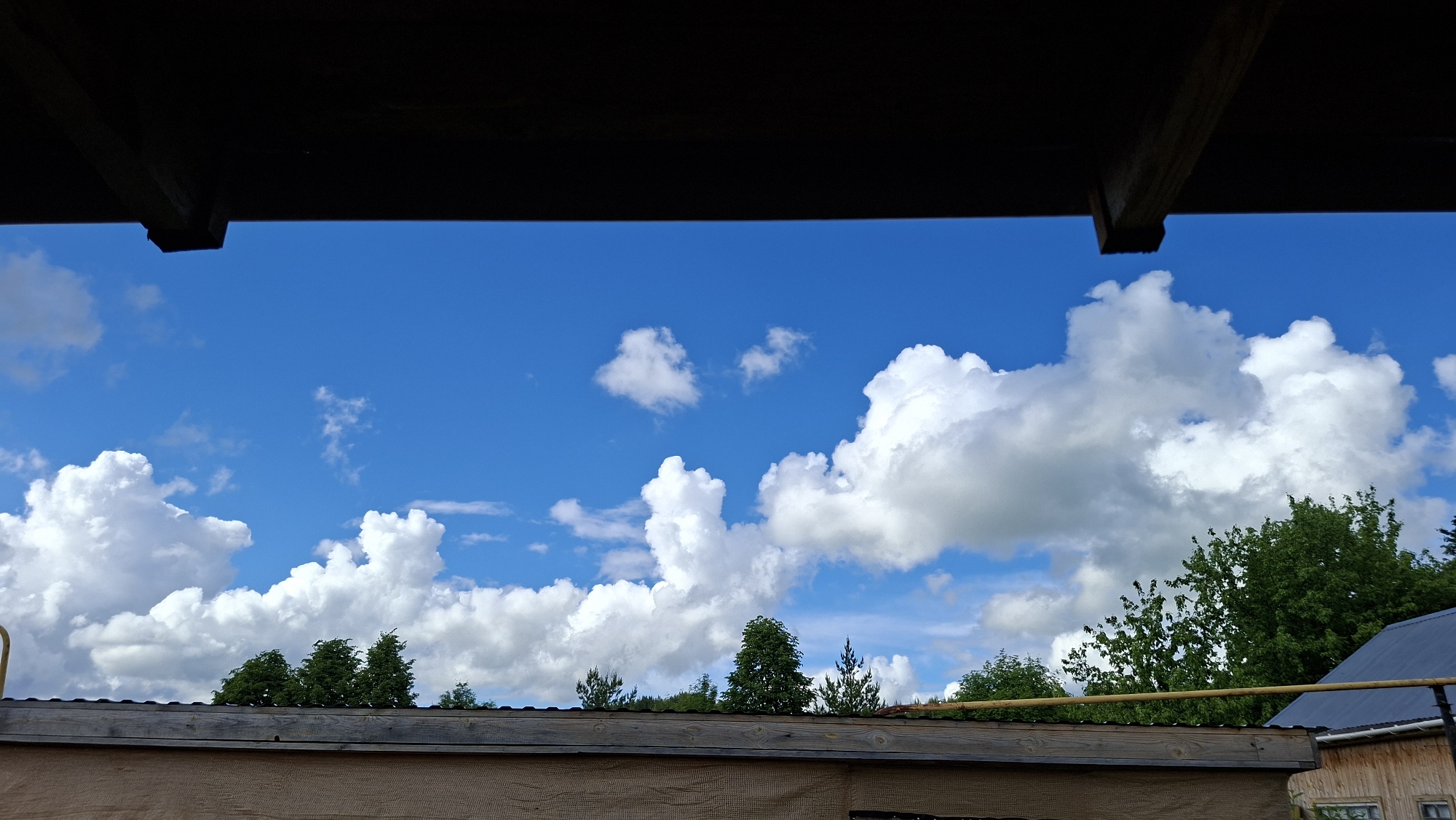 The image displays a bright blue sky adorned with numerous white, fluffy cumulus clouds of varying sizes. The scene is viewed from beneath a dark, shadowy wooden structure, likely an eave or roof overhang, which occupies the top section of the frame and shows visible wooden beams.
Across the bottom of the image, a lower wooden roofline or building structure is visible, featuring a wooden fascia board and a corrugated metal roof above it. Behind this, a mix of green deciduous and coniferous trees rise, their canopies reaching towards the sky and clouds. On the far right, a portion of another building with wooden siding and a dark gray or blue sloped roof is visible. A bright yellow pipe or rail extends horizontally in the mid-right area, positioned above the lower roofline and in front of some trees.
The time of day appears to be bright daytime, likely late morning or afternoon, given the clear blue sky and well-illuminated clouds. The weather is fair and pleasant, with no indication of precipitation. There are no people or specific activities taking place. No visible text is present. The setting, characterized by the architectural styles and natural elements, suggests a rural or semi-rural environment in Большой Букор, Russia.