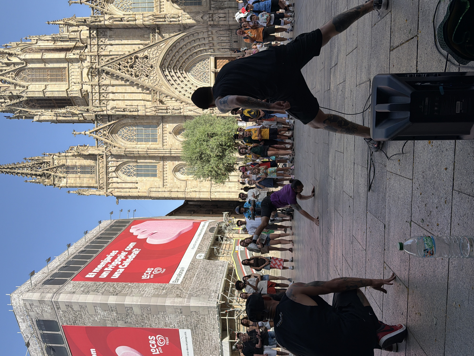 In front of the Barcelona Cathedral, two breakdancers perform for a crowd. One dancer is on the ground, his body contorted in a challenging pose, while the other is crouched, ready to join the routine. A portable speaker sits on the ground nearby, its wires trailing across the cobblestone plaza. Behind them, a large billboard advertises "Locos por Frigo," an ice cream brand. Spectators of all ages gather to watch the performance, some holding phones to capture the moment. The sun shines brightly, casting long shadows on the plaza, and the ornate Gothic architecture of the cathedral provides a striking backdrop.