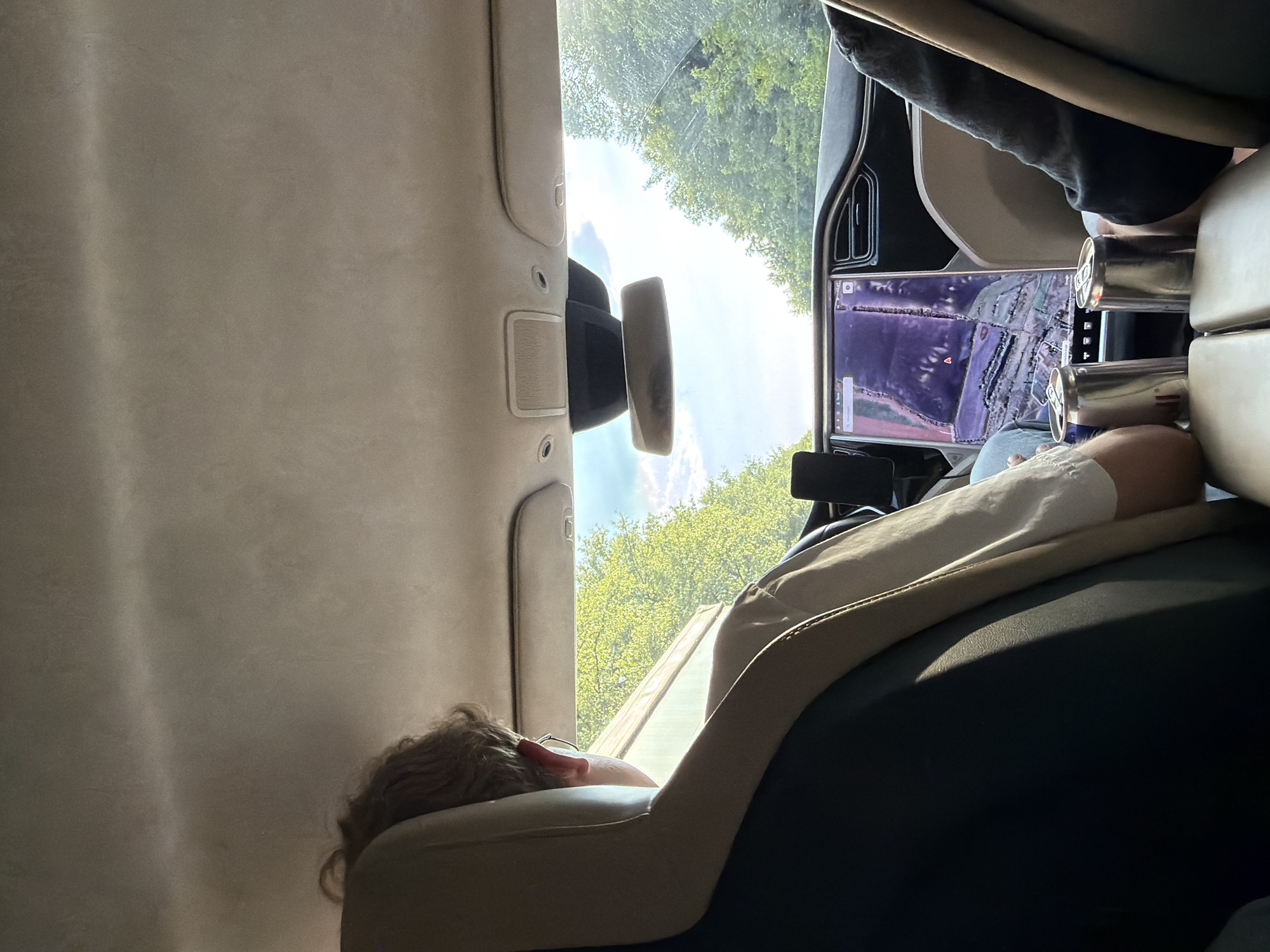 A person is resting in the back seat of a car, which appears to be a Tesla, as indicated by the large touchscreen display on the dashboard showing navigation. The person is lying down, with their head propped on a pillow and their arm resting across their lap. Their eyes are closed, suggesting they are asleep or resting. Sunlight streams into the car through the windows and sunroof, illuminating the interior and casting shadows. The trees outside suggest a rural or suburban setting with lush greenery, consistent with the location of Volytsia, Ukraine, during warmer months. The time of day appears to be midday, given the bright sunlight. The overall mood is relaxed and peaceful, with the person enjoying a comfortable rest during a car journey. Visible text includes navigation information on the car's display, but it is not clear enough to read.