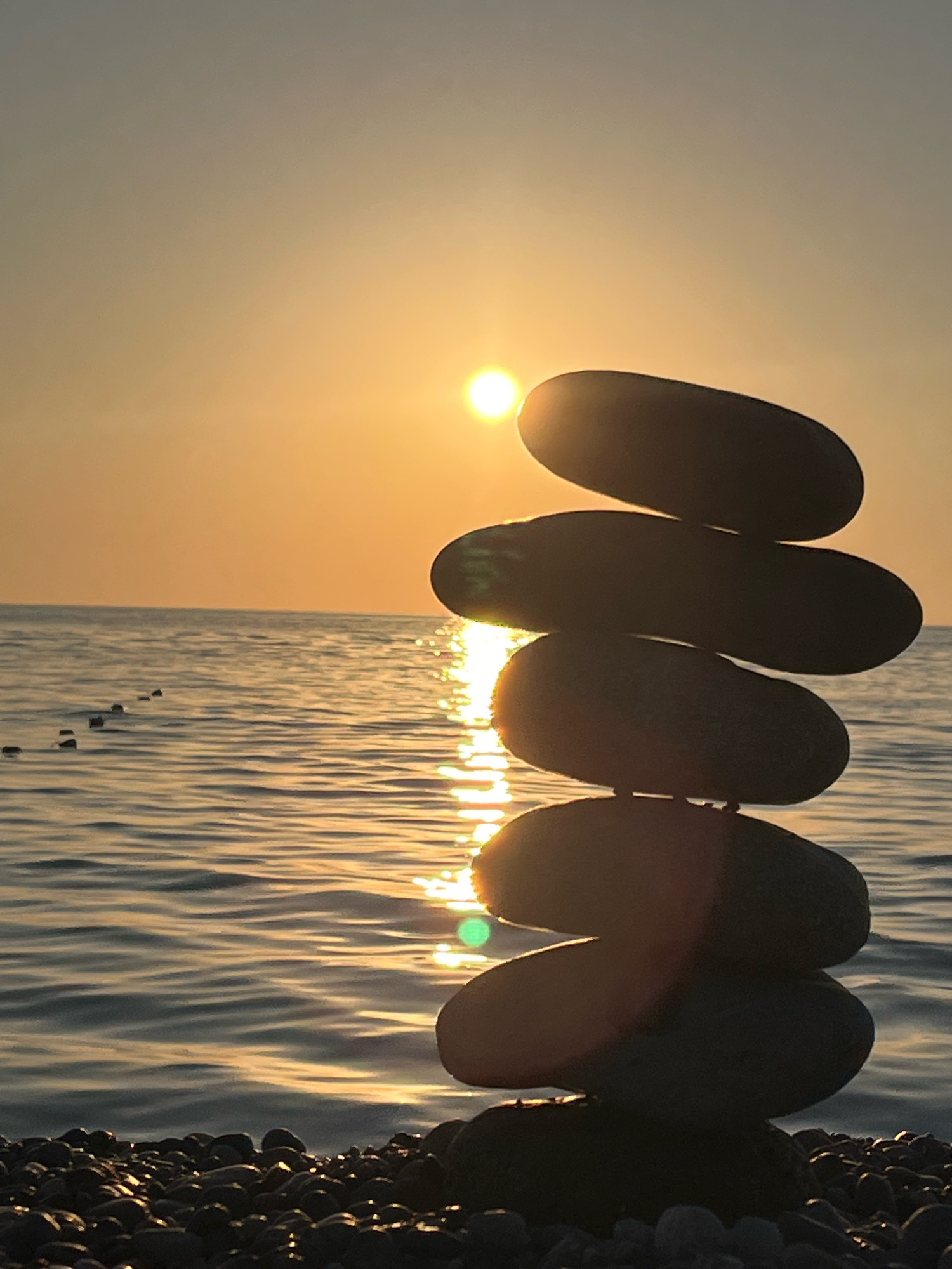 A close-up, low-angle shot captures a stack of smooth, dark stones balanced precariously on a pebbled beach, likely on the Black Sea coast of Kobuleti, Georgia. The setting sun casts a warm, golden glow across the scene, illuminating the rippling water and creating a vibrant reflection that stretches towards the horizon. The sky is a soft gradient of orange and yellow, with the sun appearing as a bright, blurred orb just above the water. A line of small, dark buoys or markers can be seen in the distance, breaking the expanse of the sea. The foreground is dominated by the meticulously arranged stones, their dark silhouettes contrasting with the luminous sky and water. The overall impression is one of tranquility and natural beauty, with the stacked stones suggesting a moment of mindful contemplation against the backdrop of a breathtaking sunset.