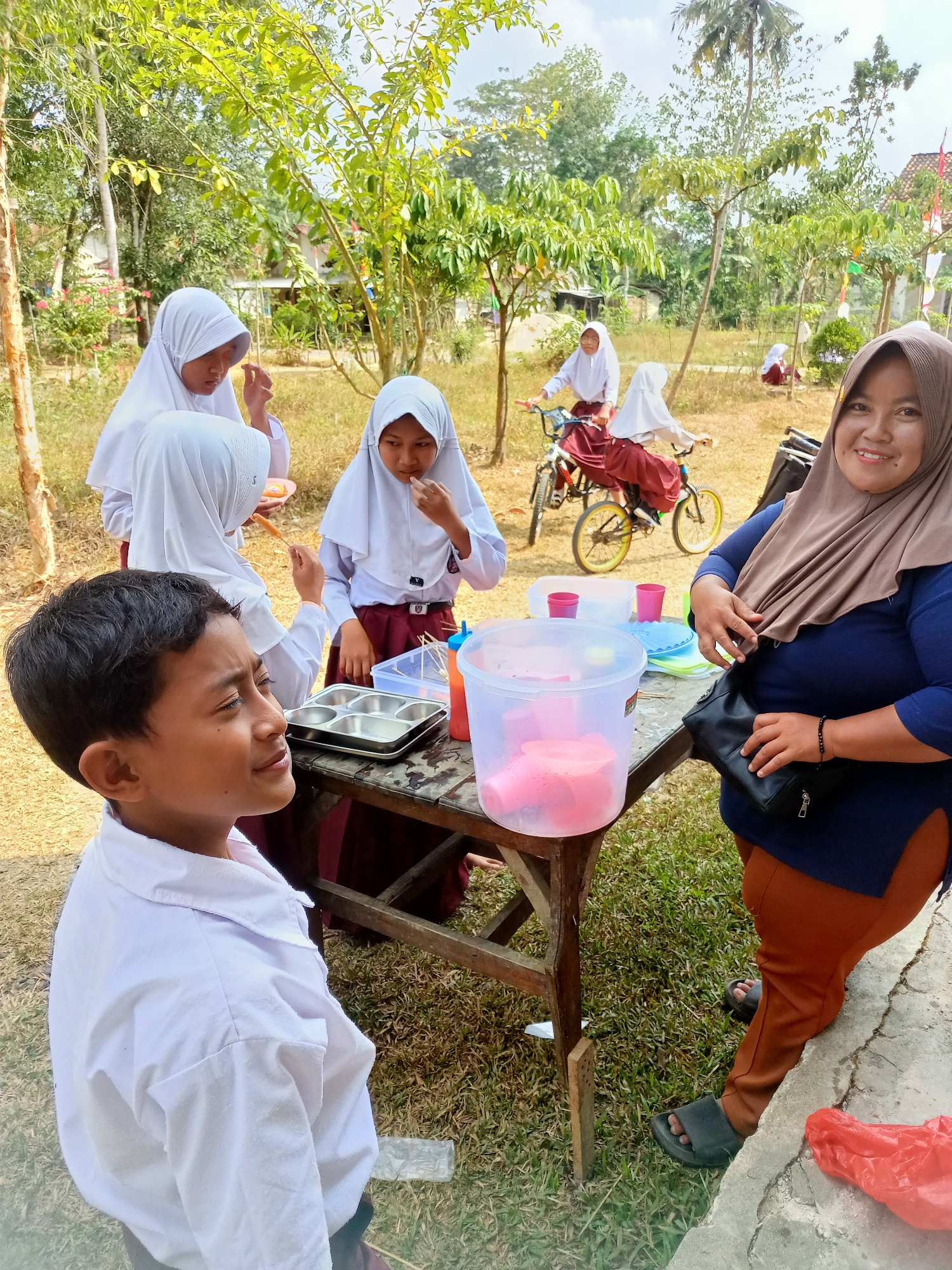 In Kagungan Ratu, Indonesia, a group of young girls in white hijabs and maroon skirts, presumably students, gather around a wooden table outdoors. One girl holds a skewer with food, while another appears to be eating. A young boy in a white shirt is positioned in the foreground, looking away from the camera. A woman in a blue shirt and brown hijab stands next to the table, smiling at the camera. On the table, there are various items including a metal tray with compartments, plastic cups, and a large clear plastic container filled with pink objects. The background shows trees, vegetation, and a house with a flag. The ground is covered with dry grass and some concrete. The scene appears to be a casual outdoor gathering, possibly during a break or an event.