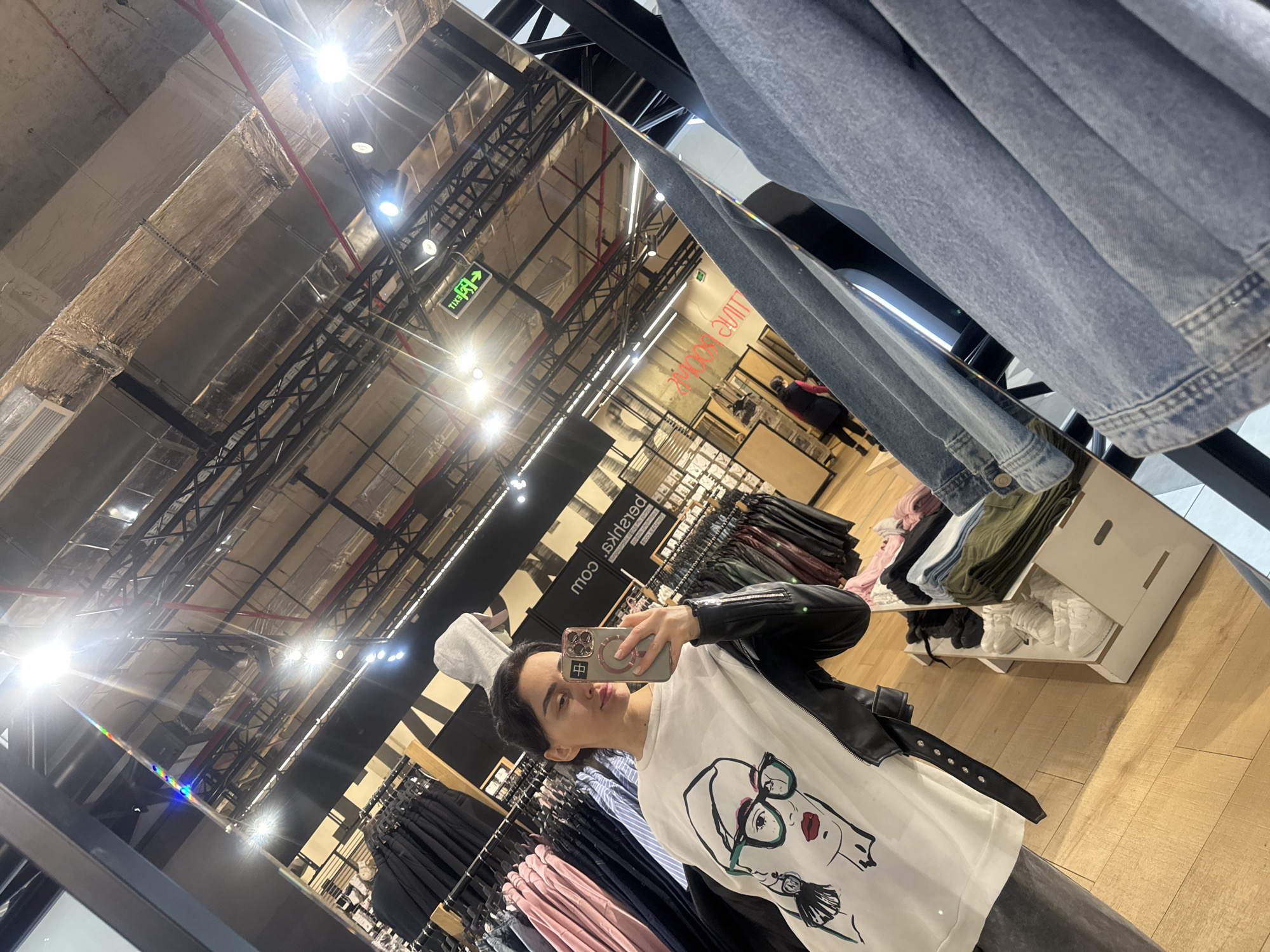 A female subject, with dark hair, is depicted in a selfie taken within a Bershka clothing store, specifically in Tbilisi, Georgia, as indicated by the reversed "Bershka.com" branding and the mirrored "EXIT" sign displaying Georgian script. The subject is wearing a white sweatshirt featuring a graphic illustration of a woman's face with green-rimmed glasses and a black leather jacket. She holds a smartphone with a multi-lens camera system and a circular magnetic accessory in her left hand, directing it towards a large, rectangular mirror.
The reflection reveals a modern retail environment with a light wooden floor and an industrial-style ceiling featuring exposed dark metal trusses, silver insulated ventilation ducts, and numerous white track lights. Clothing racks are visible, displaying various garments including black trousers, pink and blue shirts, and dark-colored jackets. Below, white display shelves hold neatly folded apparel and white sneakers. In the deeper background reflection, another individual, partially obscured, walks towards an area marked as "FITTING ROOMS." A blue denim jacket is present on a display to the right, partially framing the mirror's edge.