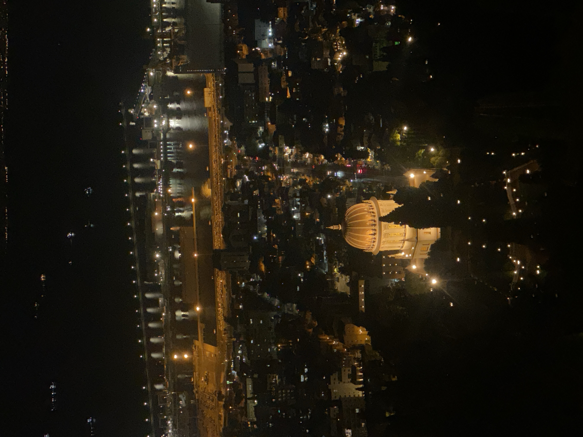 This is a nighttime aerial photograph of the city of Haifa, Israel, showcasing the Baháʼí World Centre, a prominent landmark. The image captures a wide view, with the foreground dominated by the illuminated golden dome of the Shrine of the Báb. The shrine is surrounded by dark foliage, with a pathway marked by scattered lights leading down a slope.
In the midground, the city lights stretch out, revealing a dense urban landscape with buildings and illuminated streets. A major port area is visible in the background, with its structures and ships clearly outlined by lights reflecting on the water. The harbor is active, with multiple vessels present. Further in the distance, a line of lights suggests the city extending towards the horizon. The overall scene is characterized by the contrast between the darkness of the night and the vibrant artificial illumination of the city and its structures.