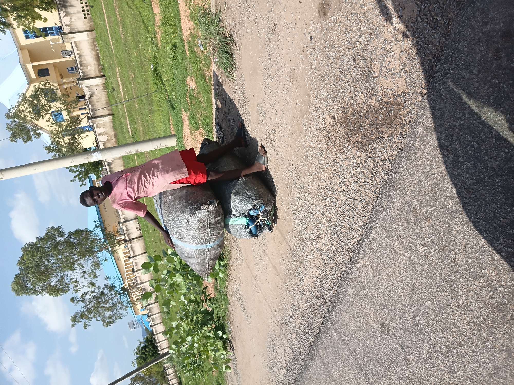 An outdoor, eye-level, medium shot shows a young Black person in a pink shirt and red shorts sitting on a pile of two large, grey, tarp-wrapped bales. The bales are on a gravelly dirt road next to an asphalt road. The person is leaning back on their hands, looking to the right. Behind them, a chain-link fence separates a grassy area from a yellow building with a blue roof. There are trees and a water tower in the background. The scene appears to be in Jalingo, Nigeria, based on the architecture and vegetation.