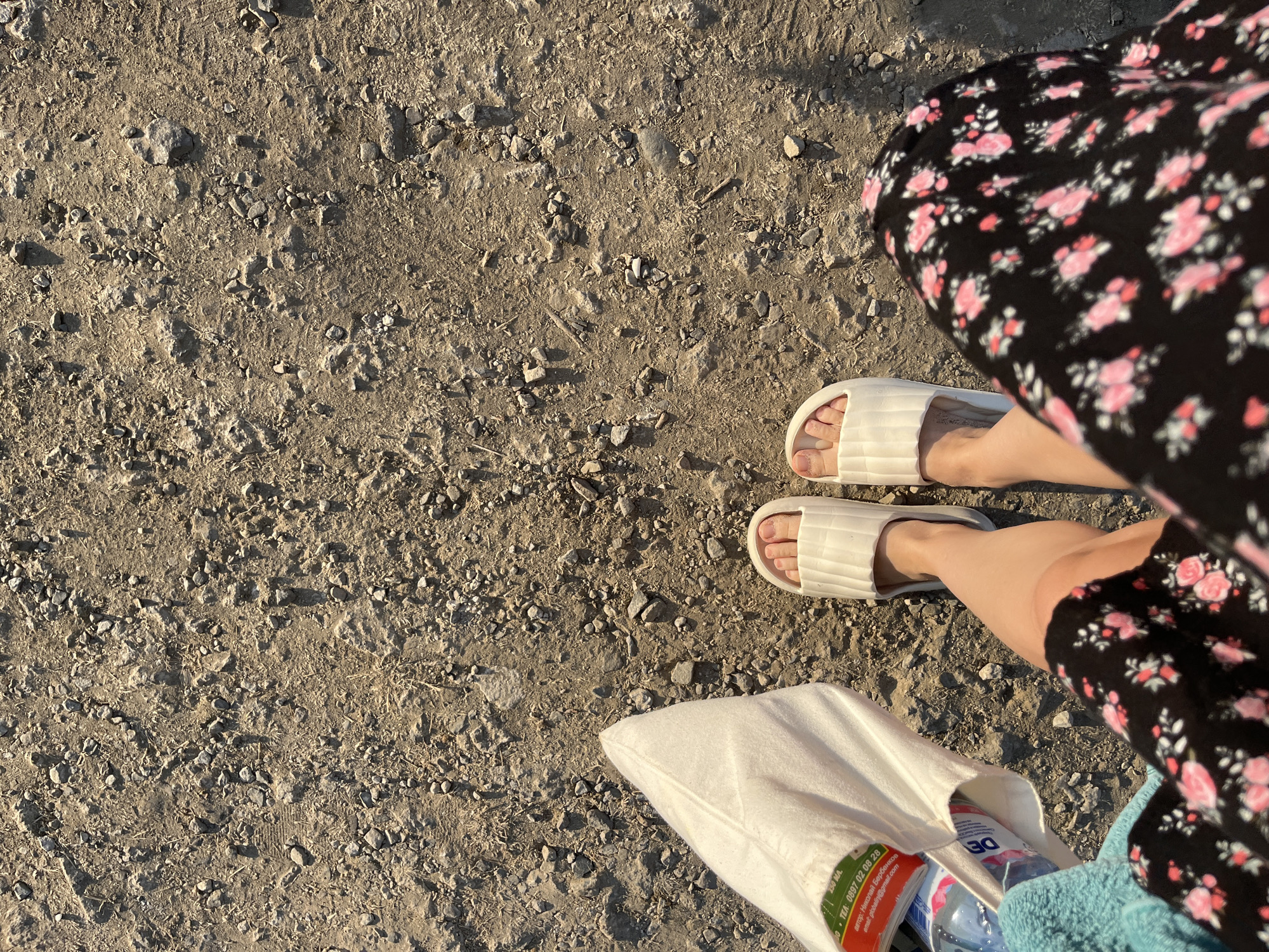 This media file shows a close-up, overhead view of a person's legs and feet on a gravelly path, with a black floral dress and white slides visible. A light-colored tote bag rests on the ground beside their feet, containing a bottle of water and other items. The scene suggests a casual outing in a dry, outdoor environment. The lighting indicates it is likely daytime, possibly late afternoon or early morning due to the long shadows cast by the gravel. The setting appears to be a path or unpaved area, consistent with a rural or coastal location like Ahtopol, Bulgaria, as suggested. There is visible text on a label within the tote bag, which reads in part: "тел: 0897 02 06 28" and "email: gpobel@gmail.com". The overall mood is relaxed and unhurried.