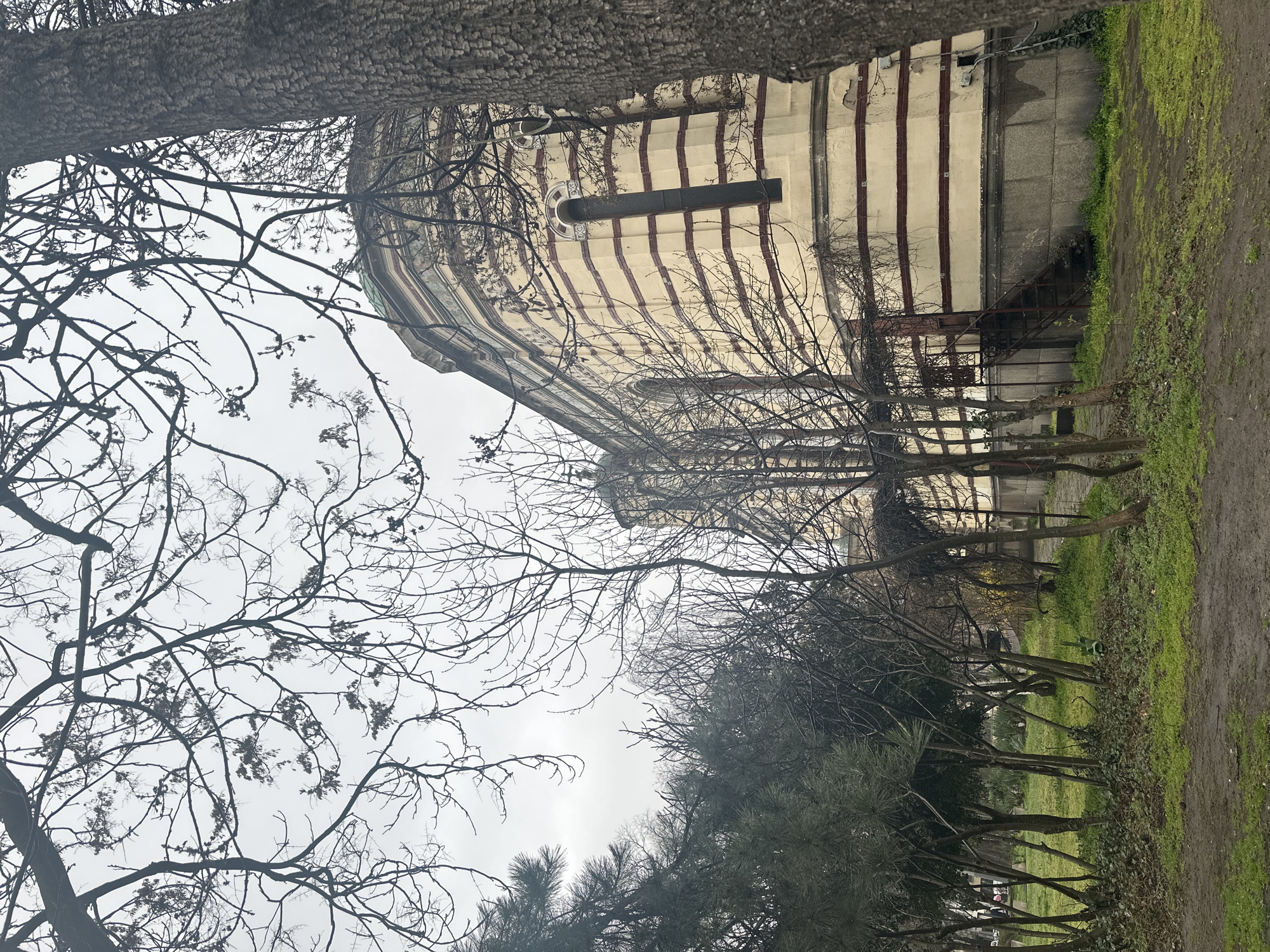 The image depicts a three-story cylindrical building with cream-colored walls and horizontal dark red stripes in a park in Sofia, Bulgaria. A grey sky is visible above and behind the trees. The building features a small arched window. A small staircase leads to the building's entrance. Several bare trees are present in the foreground and frame the building. The ground is covered in green grass and brown dirt. A large tree trunk occupies the top of the frame, obscuring part of the sky.