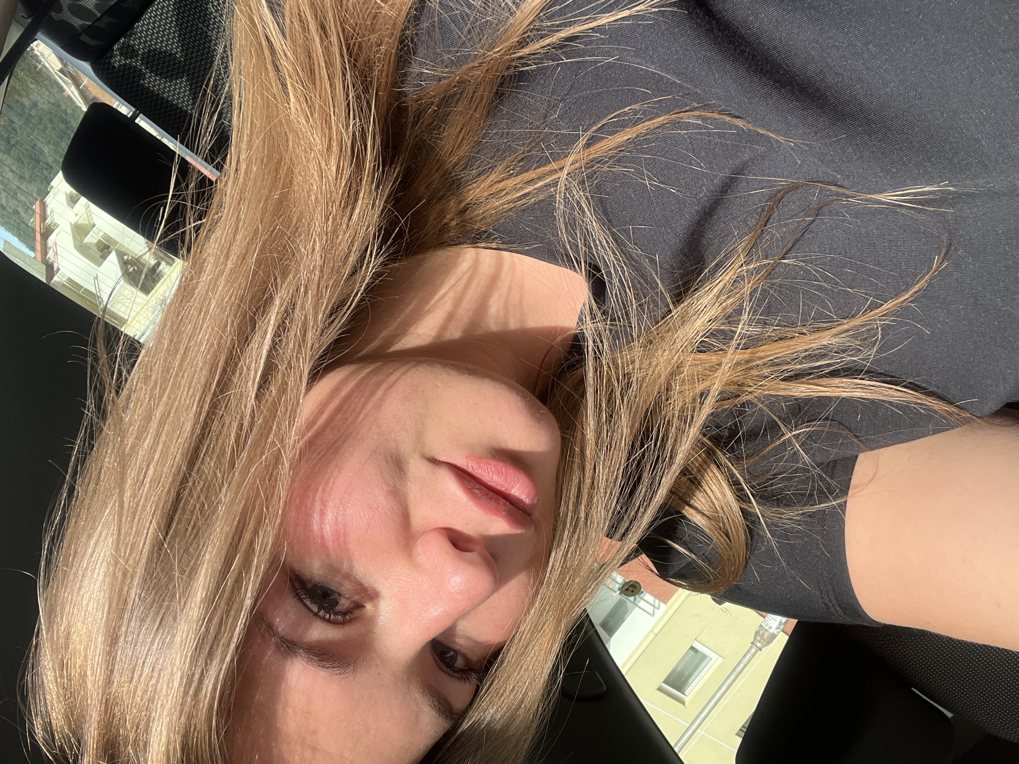 A woman with blonde hair and light skin is taking a selfie while sitting in a car. She is wearing a black shirt. The background visible through the car windows shows buildings, possibly in an urban area. The location is Budva, Montenegro based on the building color palette visible through the window.