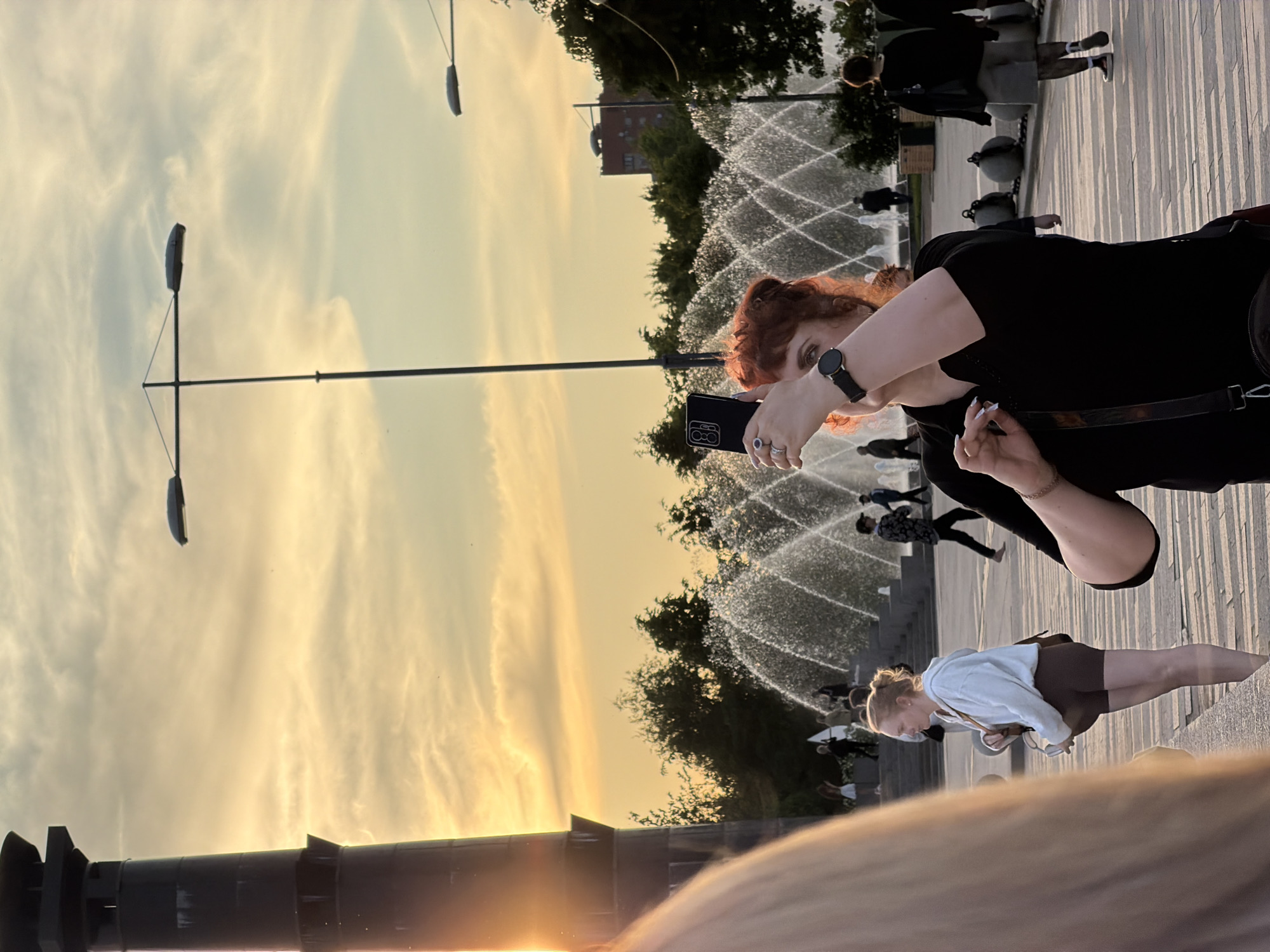 This image captures an outdoor public space in Saint Petersburg, Russia, during the golden hour, indicated by the warm, soft yellow and orange light bathing the sky and scene. The sky displays delicate, wispy cloud formations.
In the mid-ground, a woman with reddish-brown hair, wearing a black short-sleeved top and a black smartwatch on her left wrist, is the primary subject. She holds a black smartphone in her right hand, adorned with rings, and appears to be actively taking a photo or video.
Behind her, a prominent feature is a large, active fountain system with multiple water jets arcing upwards. Several other people are visible throughout the scene: one woman in a white hoodie and dark shorts stands near the fountain, looking downwards. Other figures are scattered in the mid-ground and background, some interacting with the fountain, while others walk along the paved ground, which features a distinct linear, striped pattern.
Tall, modern streetlights with multiple arms extend into the sky. Green trees line the background, with glimpses of distant buildings. The atmosphere is calm and pleasant, suggesting people are enjoying a leisurely moment in an urban park. No discernible text is visible in the image.