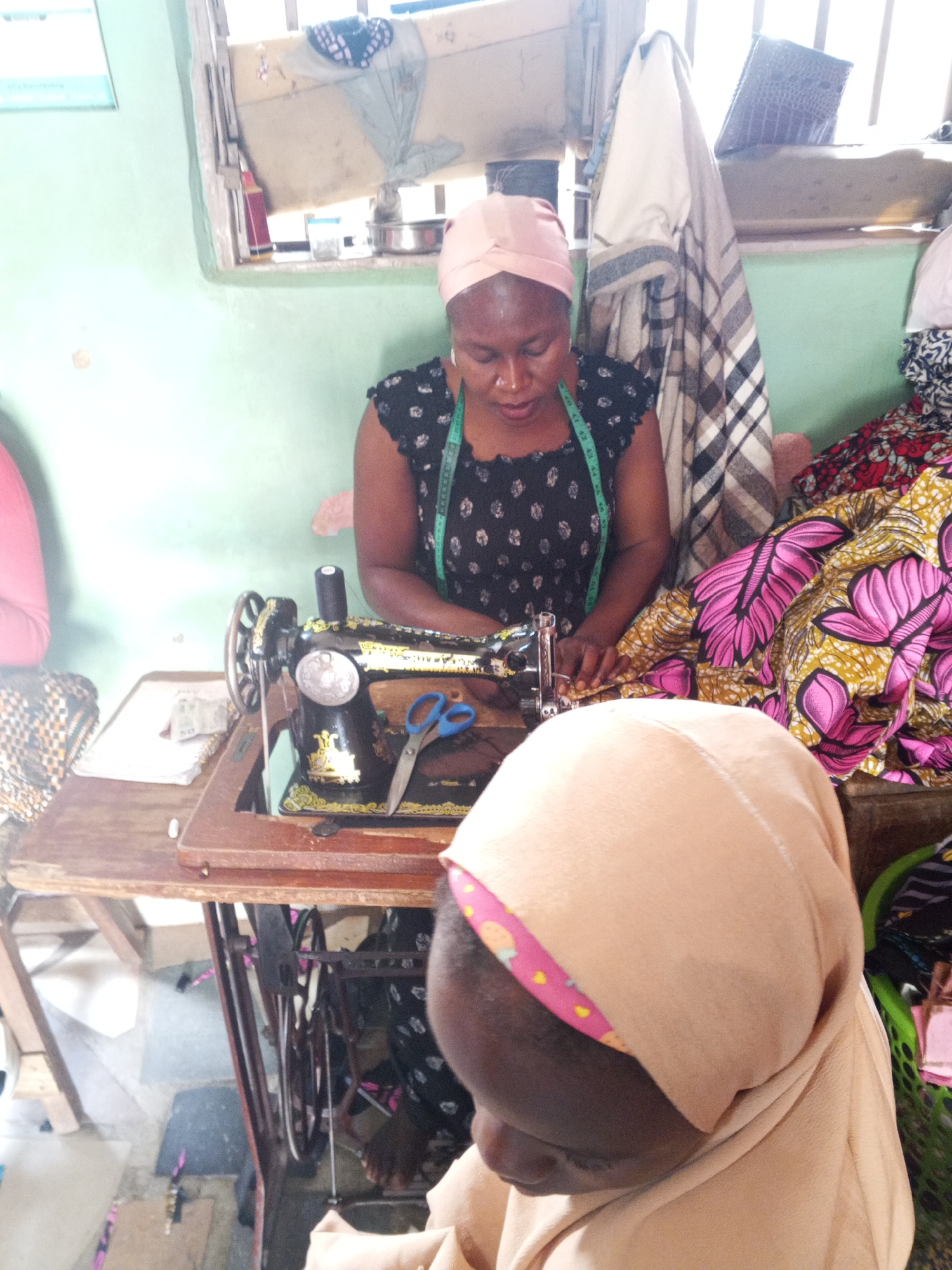 An indoor scene in Unknown, Unknown, shows a woman operating a manual sewing machine on a wooden table. The woman, positioned centrally, wears a light pink headscarf (turban style) and a dark patterned short-sleeved top with a green measuring tape draped around her neck. Her gaze is directed downwards at the fabric under the needle of the black sewing machine, which features gold embellishments. A pair of blue-handled scissors rests on the machine's table, and a black spool of thread is visible on the machine's spindle. To the woman's right, a substantial pile of vibrant patterned fabric, predominantly pink, yellow, and green, is stacked.
In the immediate foreground, the back of another person's head and left shoulder are visible, facing away from the viewer. This individual wears a light beige head covering with a pink patterned headband showing underneath.
The background features a light green painted wall. Behind the main woman, a rectangular window provides bright natural light, revealing indistinct objects on a ledge, including bottles and containers. To the right of the window, a plaid fabric item is draped, and a dark, textured object (possibly a bag) is visible above it. Further right, a plain white garment hangs. On the left wall, a white board with faint blue text is affixed. The floor, partially visible beneath the table, appears to be a light-colored, possibly concrete surface.