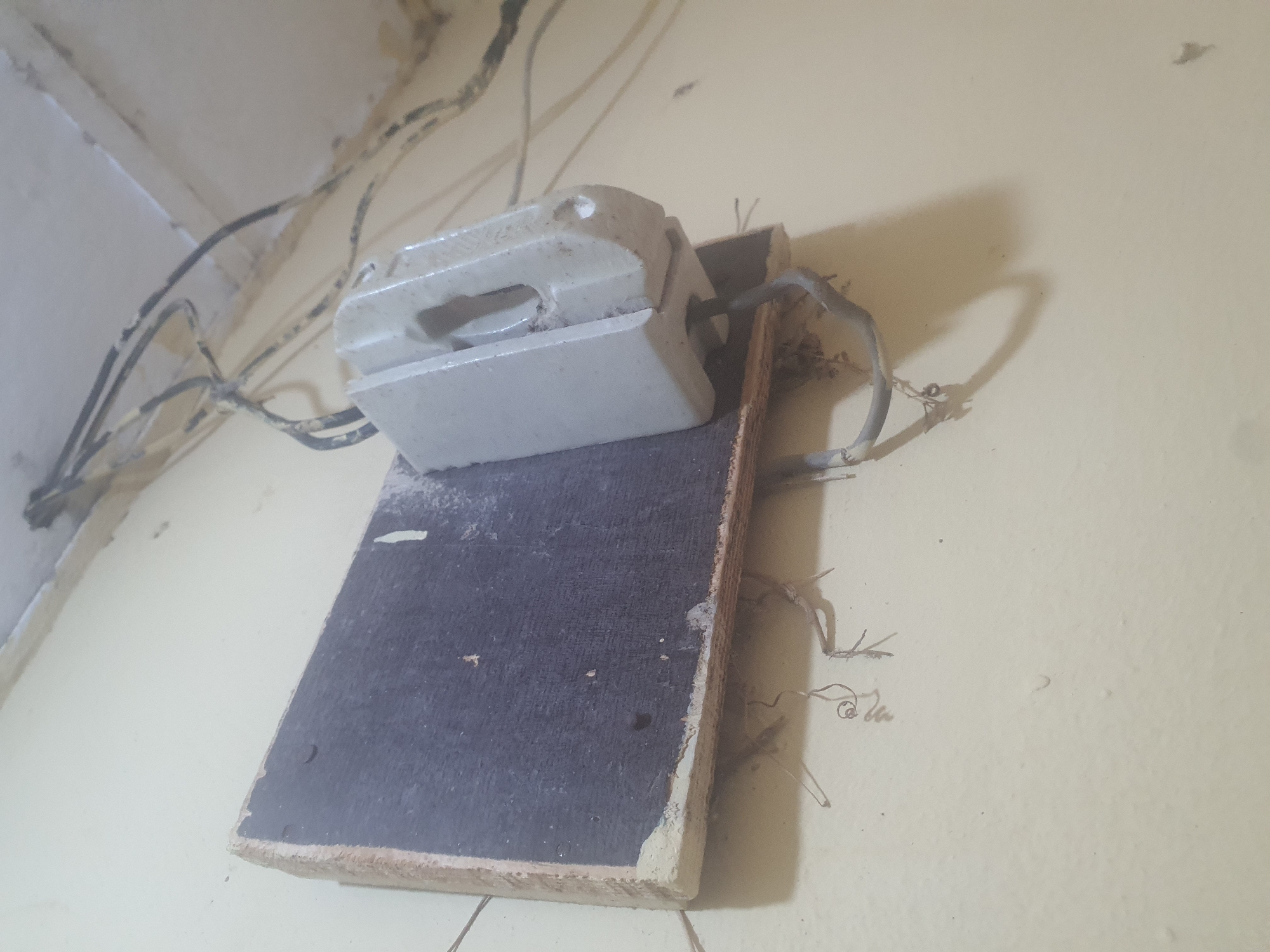 A close-up, low-angle shot shows a rudimentary electrical fuse holder mounted on a piece of wood attached to a cream-colored wall. The fuse holder is a light gray, rectangular ceramic device with a slot on top and a wider opening at the bottom where wires enter. It appears dusty and aged. The wooden backing is rough, with a dark surface on the front where the fuse holder is attached. Several electrical wires, black and gray, are visible, some frayed and exposed, snaking across the top of the frame and down towards the fuse holder. Some wires hang loosely, with visible twists and connections. The wall surface exhibits some dirt and staining, particularly near the ceiling and around the electrical installation. The environment suggests an indoor setting, likely an older or unkempt building.