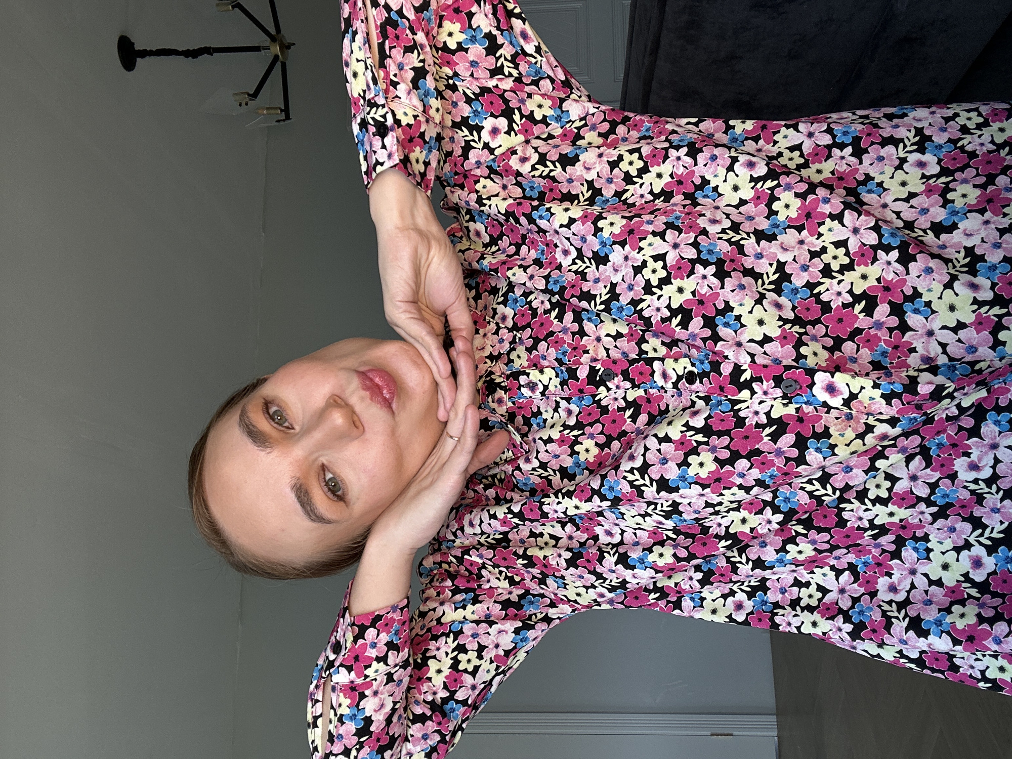 A woman with fair skin, light brown hair pulled back, and green eyes is wearing a floral-patterned dress. She has her hands positioned near her face, and she is looking towards the camera. The background includes a gray wall, a chandelier, and a dark-colored sofa or cushion. There's no indication of Kharnavy, Russia in the image.