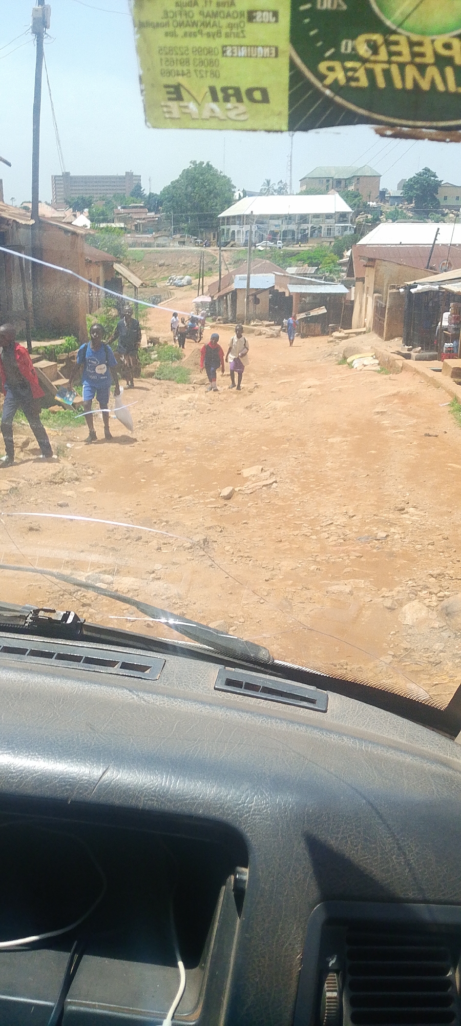 The image shows a dirt road in what appears to be a residential area. There are several people walking on the road, some carrying bags. Buildings line the sides of the road, with some appearing to be shops. The overall impression is one of a bustling, everyday scene in a developing urban environment. The text on the green sticker at the top of the image indicates that this is likely in Jos, Nigeria, as it contains the phrase "SPEED LIMITER" and "DRIVE SAFE," which are common safety slogans.