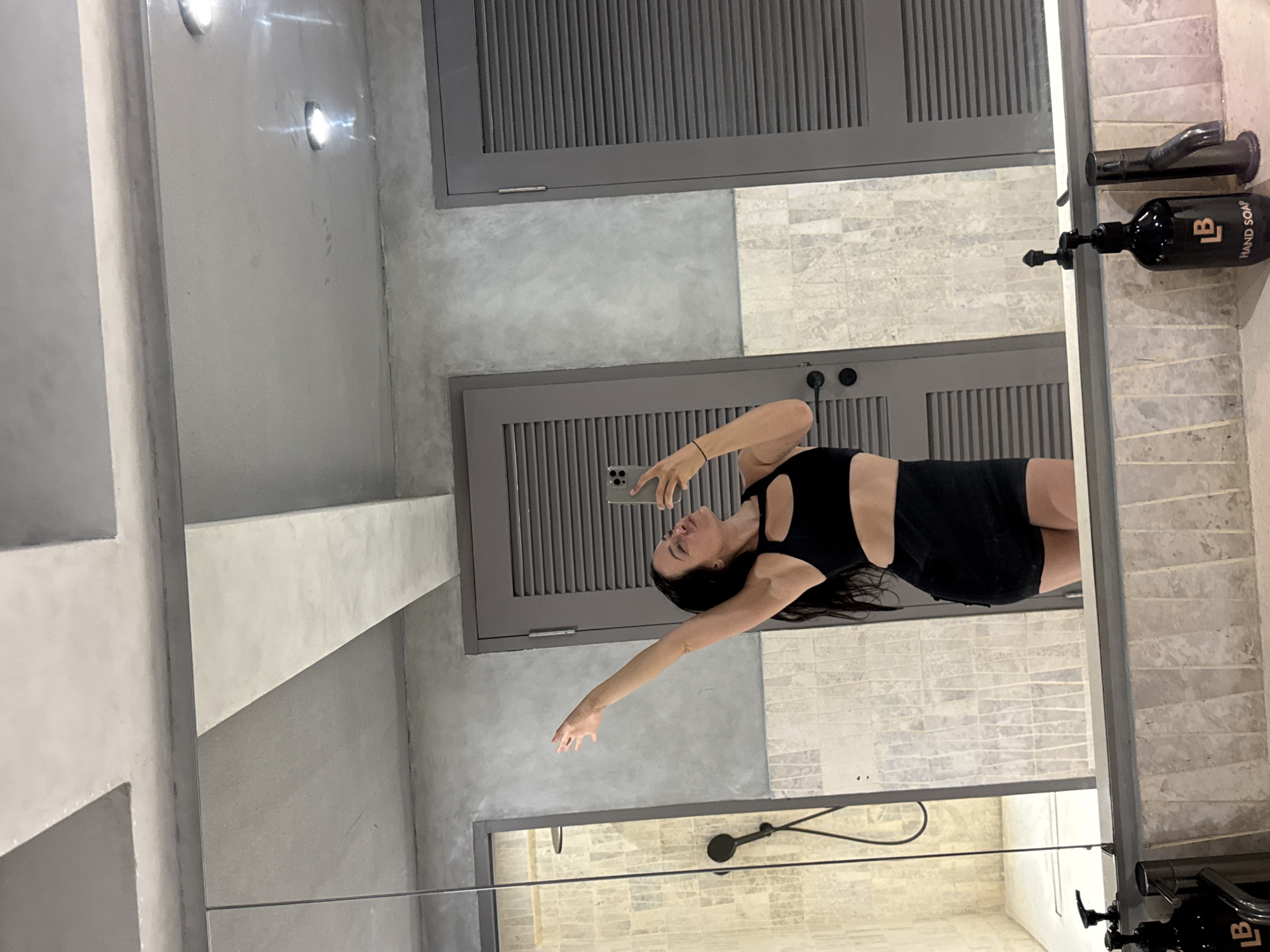 A woman with dark hair is taking a mirror selfie in a bathroom. She is wearing a black sports bra and black shorts. Her left arm is raised with her hand outstretched. The bathroom has a gray, concrete-like textured wall, a tiled shower stall, and two gray doors with louvers. There is a mirror above a vanity with a sink and two soap dispensers. The lighting in the bathroom is dim with two ceiling lights visible. This scene is in North Kuta, Indonesia.