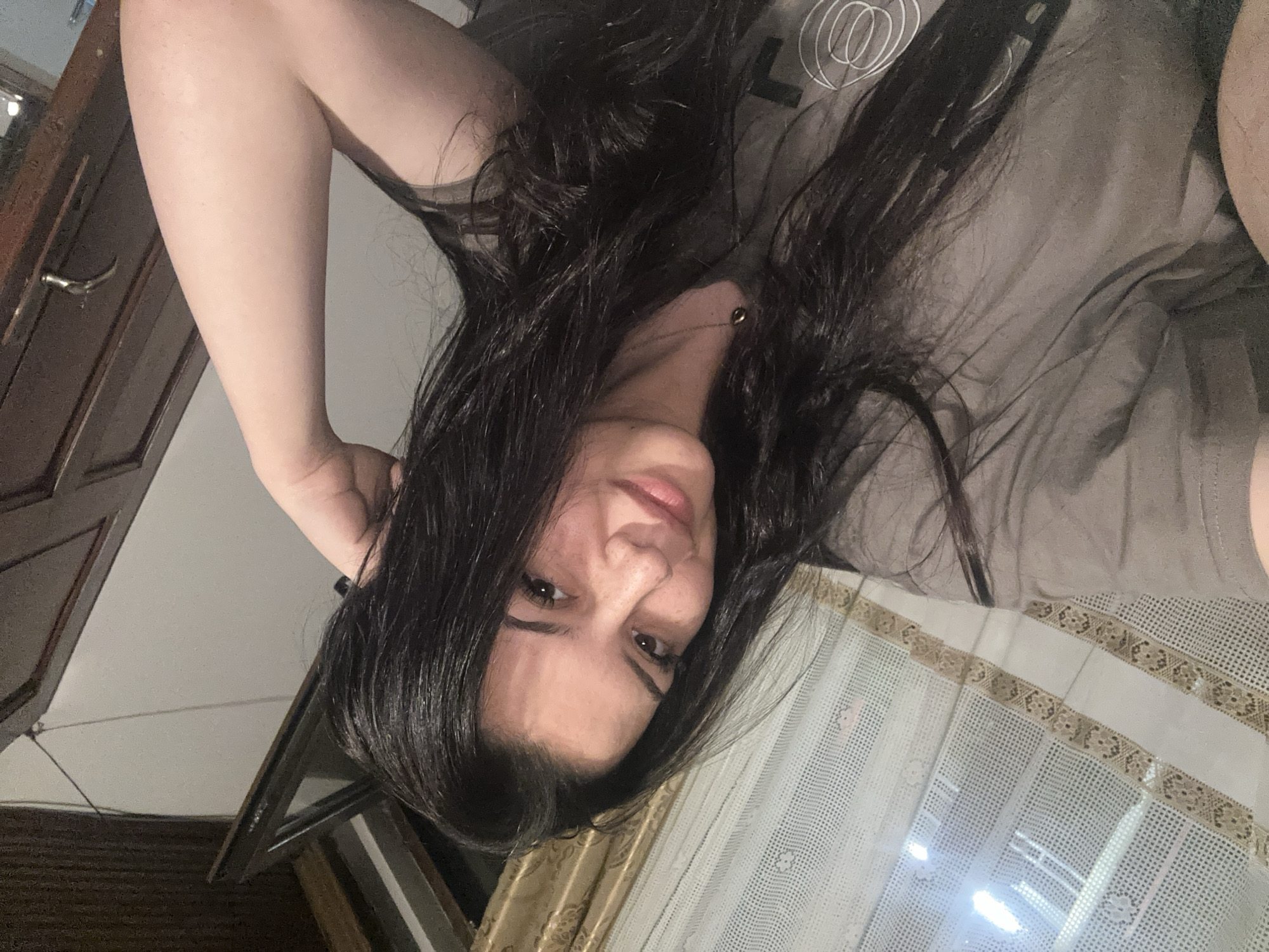 A sideways-oriented portrait captures a woman indoors, likely in Chubra, Bulgaria. The subject, facing forward, has long, dark, wavy hair and dark eyes, looking directly at the camera with a neutral to slightly smiling expression. Her nose and cheeks show visible freckles. She is wearing a grey t-shirt with a white graphic design of concentric circles on the left side, and a thin, gold-colored necklace with a small pendant. Her left arm is bent, with her hand positioned near her head and hair.
The background indicates a home interior. To the left, a dark wooden door or cabinet with a metallic handle is partially visible. To the right and below, a window is dressed with two layers of curtains: an inner white, semi-sheer curtain featuring a mesh or lace pattern with decorative horizontal bands in a brownish-gold hue, and an outer, solid light gold or beige curtain. Through the window, bright artificial light sources or reflections are visible. The overall lighting suggests an indoor setting, possibly in the evening or at night. There is no discernible text beyond the graphic on the t-shirt.
