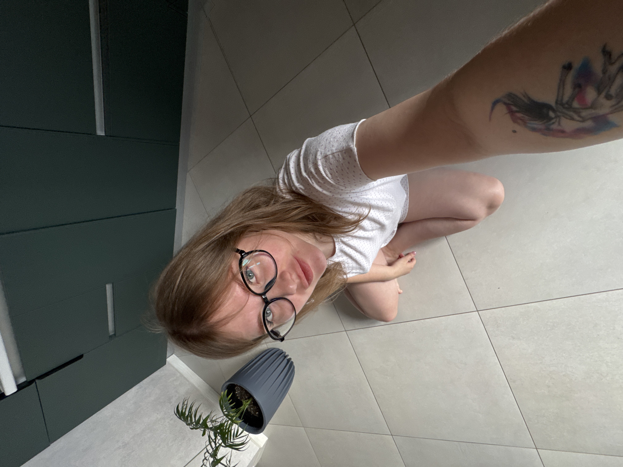 The image depicts a female individual in an indoor residential setting in Gornyi (Dvurechenskoe S/P), Russia. The subject, with light brown hair and black-rimmed eyeglasses, is positioned in a crouched or kneeling posture on a light-colored tiled floor. She is dressed in a short-sleeved white garment featuring a subtle dotted pattern. Her lower limbs are bare. A multi-colored tattoo, depicting a stylized figure, is present on her right forearm. To the left of the subject, a green plant is contained within a dark gray, fluted pot resting on the floor. In the upper background, dark green, handleless wall-mounted cabinetry, featuring a metallic horizontal accent, is visible against a light-toned wall. The subject is looking directly towards the camera, which is held at an upward angle.
