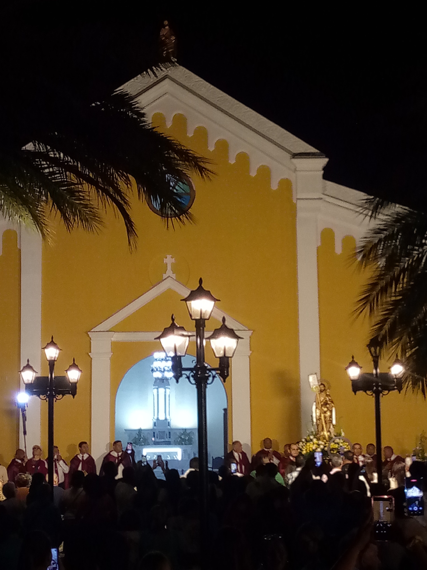 An outdoor religious gathering takes place at night in front of a church with a bright yellow facade and white trim. The church features a prominent central archway, a cross above the entrance, and a round stained-glass window on the upper left. A small statue crowns the peak of the church's roof. Palm trees frame the scene, their fronds dark against the night sky.
In the foreground, a crowd of people is gathered, their faces and details mostly obscured by the darkness, but many are seen holding up cell phones to capture the event. Several multi-branched street lamps with glowing lanterns illuminate parts of the scene.
The focal point of the event is a golden statue of a saint, possibly John the Baptist given the location context, standing on a elaborate floral base. This statue is surrounded by a group of men wearing maroon or purple robes, some with white sashes, suggesting they are clergy or part of a religious order or choir. They appear to be leading or participating in a procession or ceremony. Through the brightly lit archway of the church, an illuminated altar or interior structure is visible. The bright artificial lights create a festive and reverent atmosphere against the dark backdrop of the night. No discernible text is visible in the image.