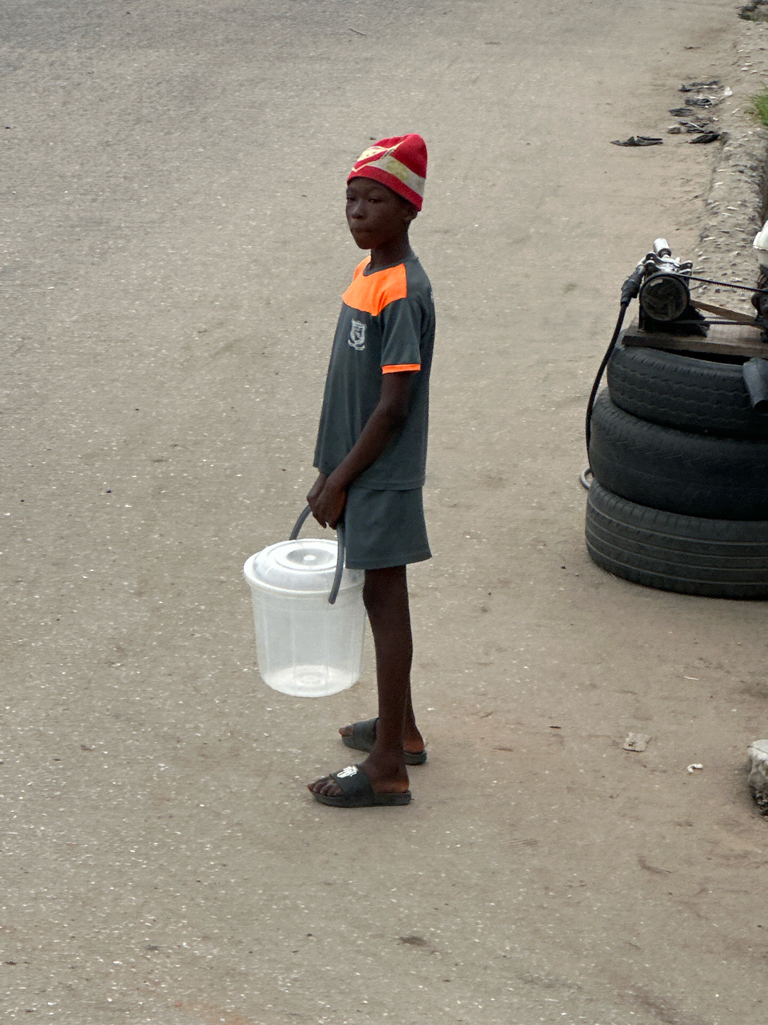 A young person, likely a child, stands on a paved surface in Ajara, Nigeria, holding a white plastic bucket with a dark handle. The child wears a red and white patterned beanie and a dark gray t-shirt with orange trim. The t-shirt has a crest visible on the left side. They are also wearing a dark gray skirt and black flip-flops. The ground is asphalt with scattered small stones and debris. To the right of the child, a stack of three black tires is visible, with a metal object, possibly a generator, placed on top of them. The background is out of focus, showing a hint of vegetation and more debris along the edge of the road.