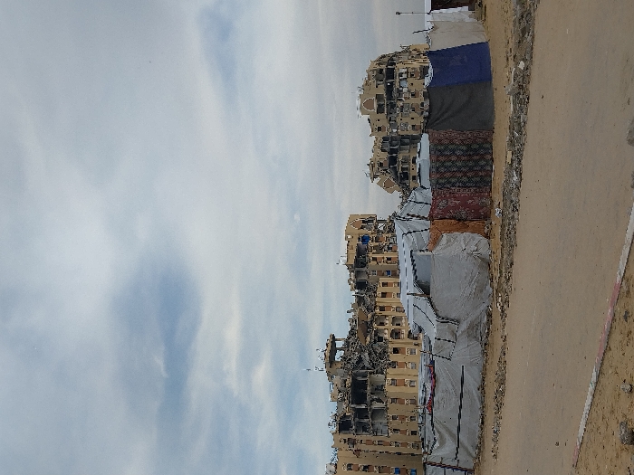 The image depicts a scene of extensive structural damage and makeshift living conditions in Khan Yunis, Palestinian Territory, under an overcast sky. In the background, two multi-story apartment buildings, constructed of light-colored concrete or stucco, exhibit severe damage. Large sections of the upper floors are missing or collapsed, exposing internal structures, rebar, and darkened interiors. Numerous windows are shattered or blown out, leaving empty frames or gaping holes. The buildings show uniform architectural features, including arched window designs and rooftop elements, now largely destroyed.
In the midground, a row of makeshift shelters or tents extends horizontally across the frame. These structures are constructed from various materials, primarily white and grey tarpaulins, with some sections made from patterned fabric, including what appears to be rugs or carpets in red, brown, and blue hues. The shelters are low-rise and appear to be temporary, with fabric draped and secured to form walls and roofs. No people are visible within or around these shelters.
The foreground consists of an unpaved, sandy or dusty ground surface. A narrow, lighter-colored strip, possibly a dirt road or path, runs horizontally across the lower third of the image, bordered by scattered debris and rubble. No vehicles or discernible human activity are present. The overall environment conveys a sense of devastation and displacement.