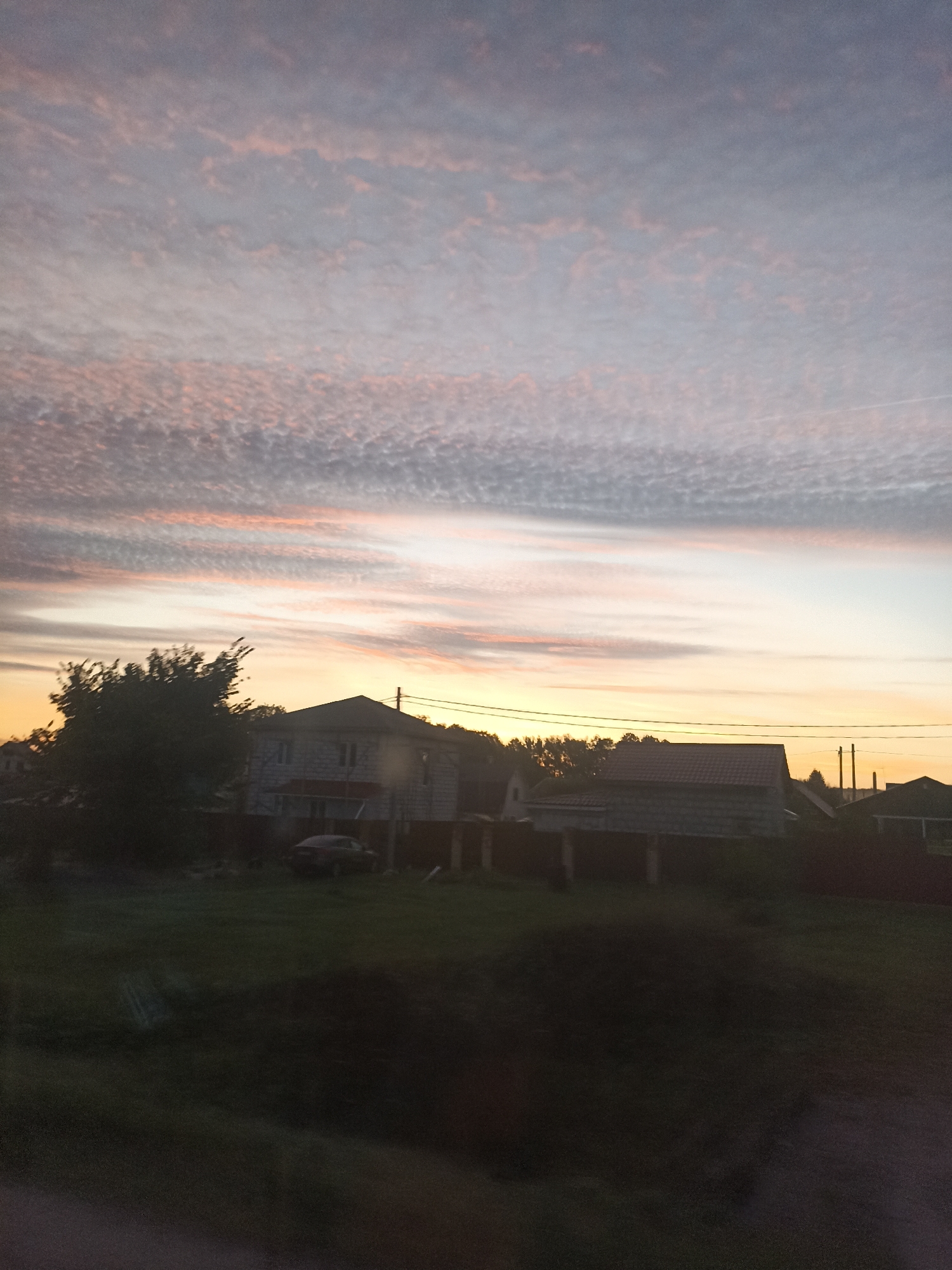 A low-angle, slightly out-of-focus photograph captures a twilight sky over a rural landscape in Volosovo, Russia. The sky is dominated by wispy clouds tinged with pink and orange hues from the setting sun, contrasting with the muted blue and gray of the upper atmosphere. Below the sky, silhouetted trees and buildings are visible.
In the mid-ground, two houses are depicted. The house on the left appears to be a multi-story dwelling with white siding and a darker roof. A dark-colored car is parked in front of it on a grassy area. To the right, another house with a darker roof and lighter walls is visible. A fence runs in front of these structures. Several utility poles with wires stretch across the scene.
The foreground is dark and indistinct, suggesting either a shadowy area or a vehicle interior from which the photograph was taken. The overall impression is of a serene evening in a residential area on the outskirts of Volosovo.