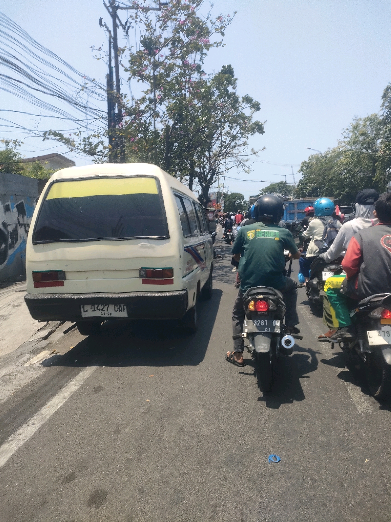 The image shows a busy street scene in Surabaya, Indonesia, likely during the day under a clear sky. A white van is parked on the left side of the road, facing away from the camera. Its license plate reads "L 1427 CAF". In front of the van, several motorcycles are seen in traffic. The riders are wearing helmets and casual clothing. The motorcyclist closest to the camera is wearing a dark green t-shirt with text on the back, and is on a motorcycle with license plate "L 3281 OW". Other motorcycles and riders are visible further down the road. The environment appears to be urban, with some trees and buildings visible in the background, along with power lines overhead. The road surface is asphalt and appears to be in good condition.
