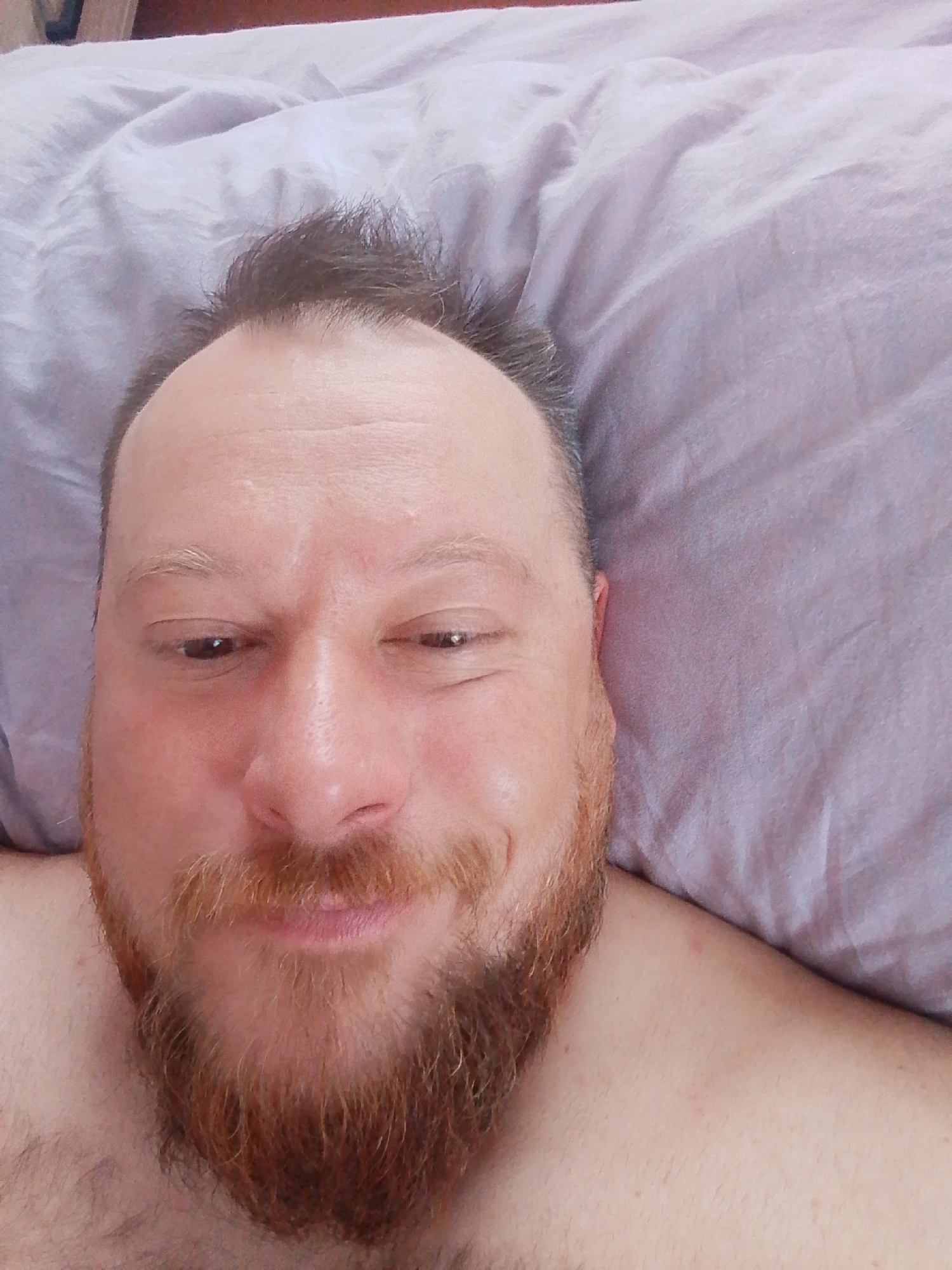 A man is lying in bed. He has brown hair, hazel eyes, and a red beard. He is smiling slightly. The background is a light purple pillow. The location is not discernable from this image.