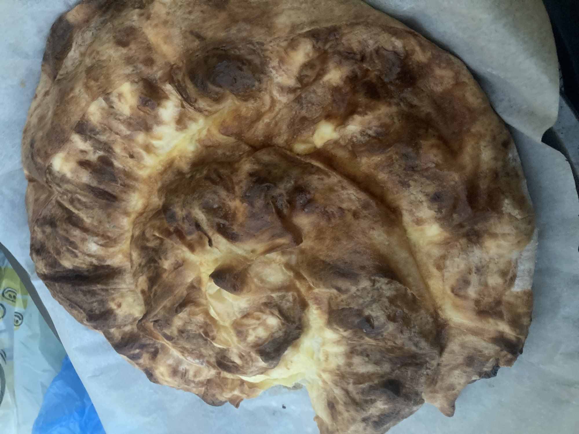 A large, golden-brown pastry with a swirled, layered appearance is presented on a sheet of white parchment paper. The pastry exhibits areas of darker browning, indicating it has been baked. The background shows a blurred blue plastic bag and a sliver of white packaging with a faint graphic. The scene appears to be indoors, possibly a kitchen or a food preparation area, but no specific location in Burgas, Bulgaria can be determined from the visual information.