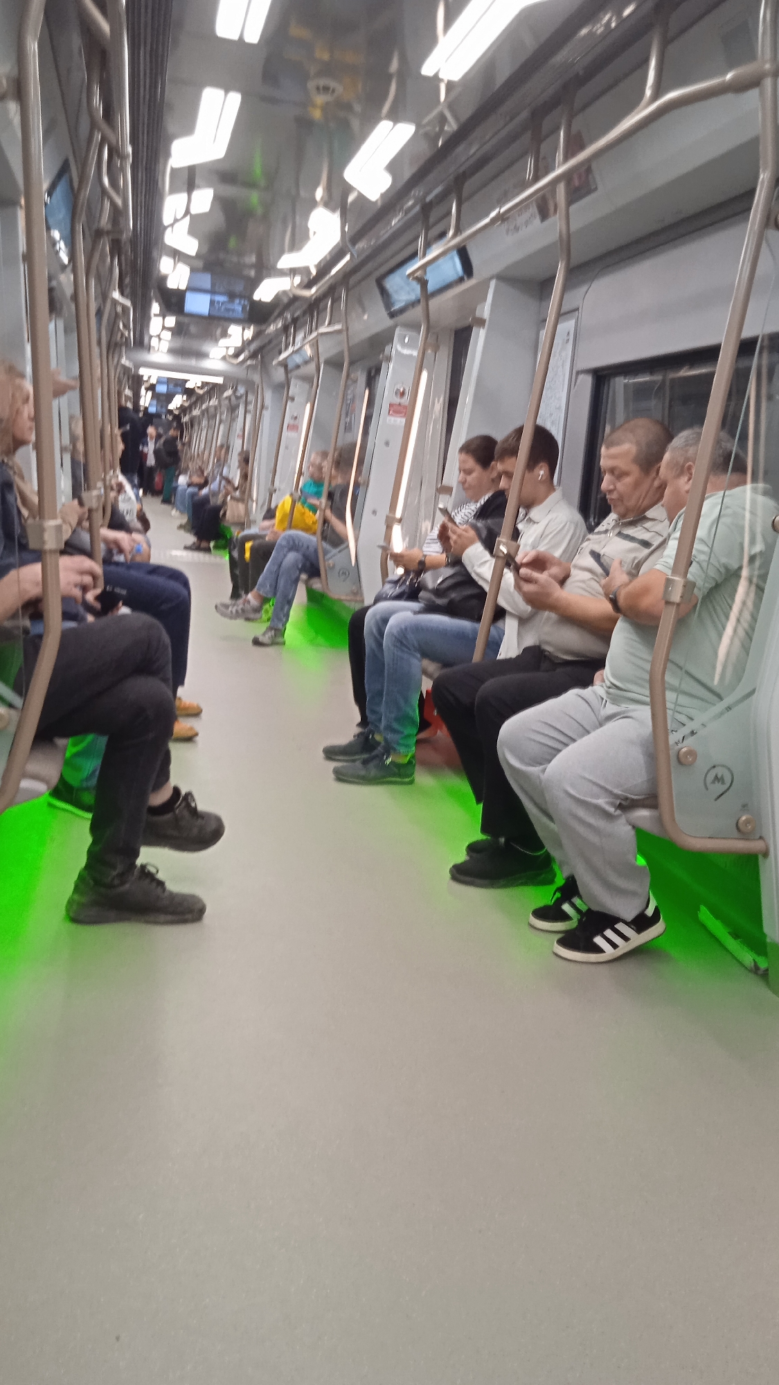 An eye-level shot captures the interior of a subway car with passengers seated and standing. The floor of the subway car is illuminated with vibrant green lighting. In the foreground, on the left, a person's legs are visible, clad in black pants and dark sneakers, with their legs crossed. To the right of the foreground, the legs of several other passengers are also visible. The passengers are seated on bench seats facing each other. Most of the passengers are looking at their phones. One person in the foreground on the right is wearing light grey pants and black sneakers with white stripes. The subway car's interior is utilitarian, with metal poles and overhead luggage racks. The overall impression is a typical scene of public transportation.