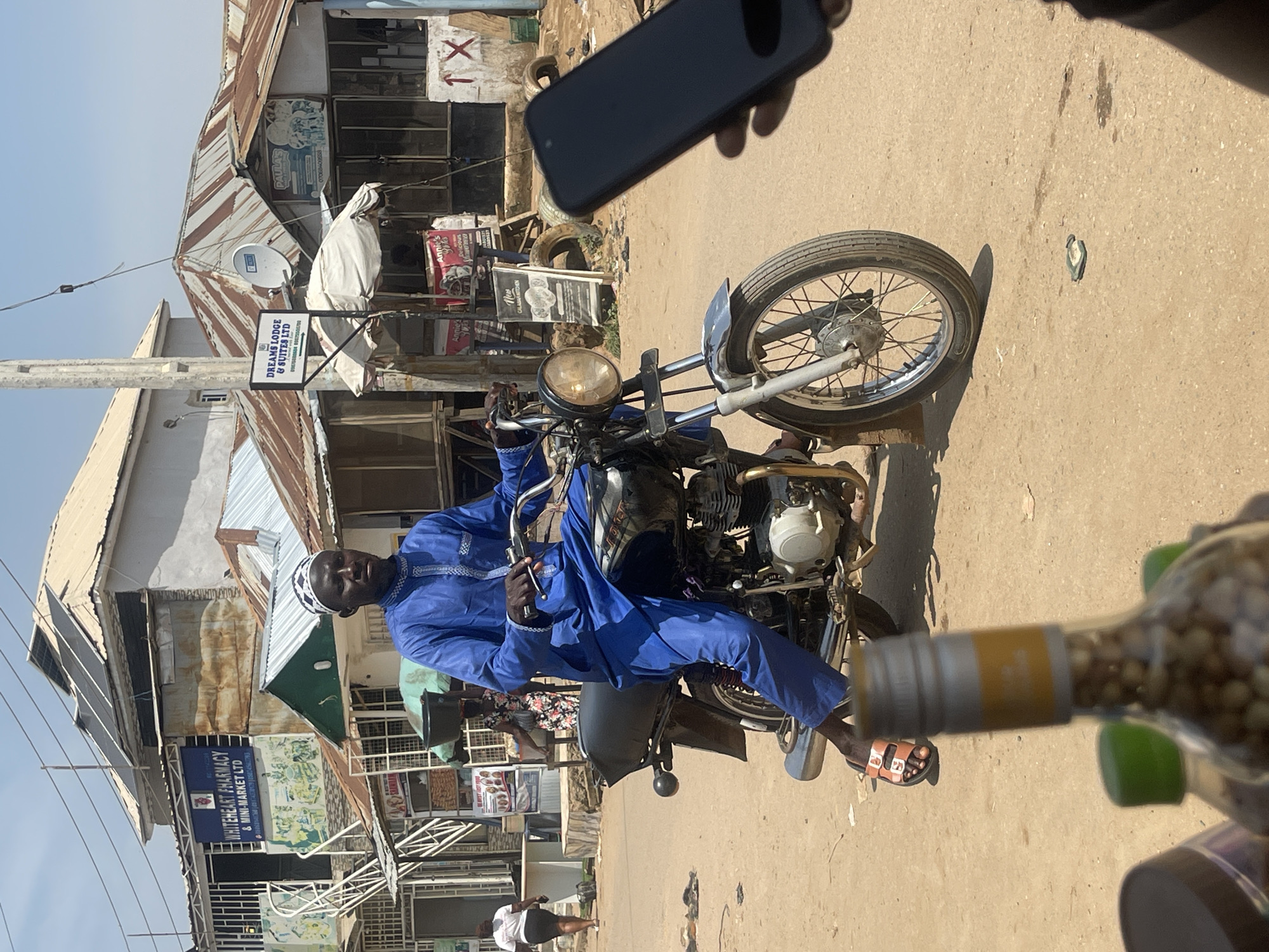 In Abuja, Nigeria, a man wearing a blue traditional outfit and a white and black patterned cap rides a black motorcycle down a dusty street. He is positioned in the center of the frame, facing forward with his hands on the handlebars. The motorcycle has a visible engine and chrome accents. Behind him, several buildings line the street, some with corrugated metal roofs and others appearing more modern. One building features a sign for "WHITEHEART PHARMACY & MINI-MARKET LTD" and another for "DREAMS LODGE & SUITES LTD." The ground is covered in loose dirt and debris. In the foreground, out of focus, are the tops of bottles, one with a yellow label. To the right, a hand holds a black smartphone, obscuring part of the scene. The sky is bright blue, suggesting a sunny day.