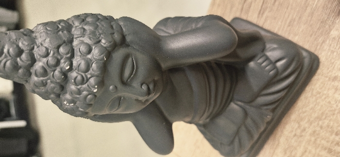 A dark grey, matte-finished statue of a Buddha-like figure is depicted seated in a meditative posture, approximately 20-30 centimeters in height. The figure's hands are raised, with palms covering the ears, and its eyes are closed. A small, circular urna mark is present on the forehead. The head features a prominent ushnisha and sculpted curls, with multiple small white scuffs or paint chips visible on the top and side surfaces. The statue is positioned on a light brown wooden surface exhibiting visible grain. The background is out of focus, revealing a lighter-toned wall or surface behind the statue and an indistinct, dark object partially visible in the upper left section. The scene is located indoors in Vendin-le-Vieil, France.
