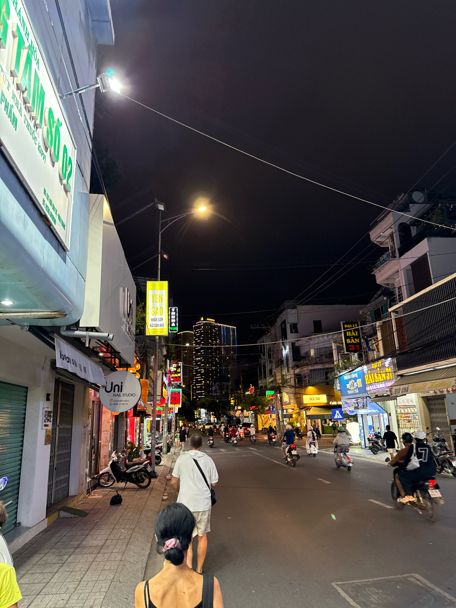 The image depicts a street scene at night in Nha Trang, Vietnam, likely in a commercial or mixed-use area. Several buildings line the street, displaying illuminated signage in Vietnamese. The architecture appears to be a mix of modern and older styles, with balconies and shopfronts visible.
Pedestrians are seen walking on the sidewalk and crossing the street. The primary mode of transportation appears to be motorcycles and scooters, with numerous vehicles on the road and parked along the sidewalk. The street is paved and appears to be well-lit by streetlights and the lights from the buildings and vehicles.
Prominent signage includes "YEN SAO" (swiftlet nest), "UNI NAIL STUDIO," and other commercial establishments. The overall atmosphere is that of a bustling urban environment at night. The sky is dark, suggesting it is late evening or early morning. The image is taken from a slightly elevated perspective, looking down the street.