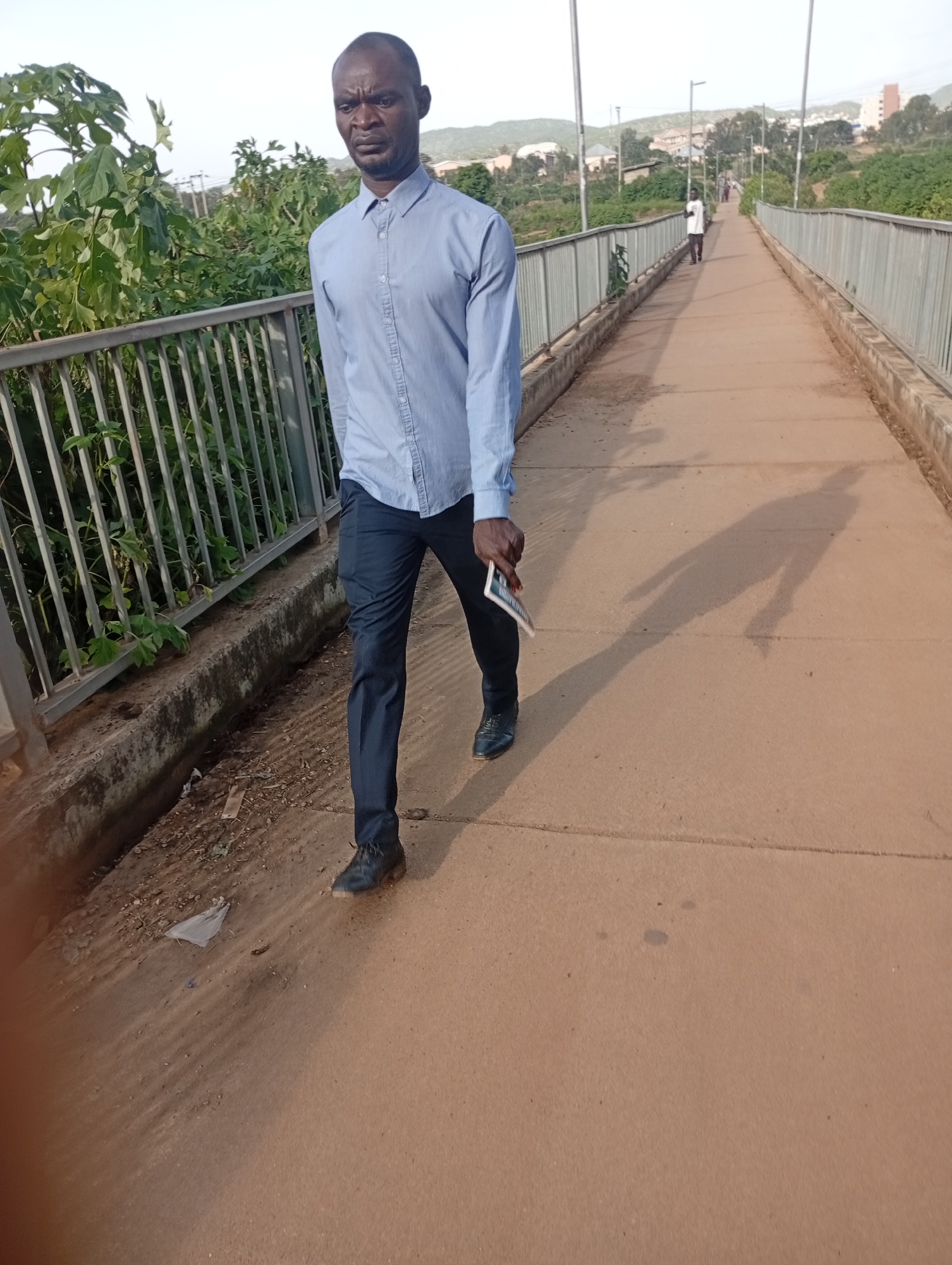 A man is walking on a pedestrian bridge in Rafin Par, Nigeria. He is wearing a light blue collared shirt and dark trousers, with black dress shoes. He is holding a small, rectangular object in his right hand. The bridge has a metal railing on the left and a concrete barrier on the right. Lush green foliage grows on the embankment to the left of the bridge. In the background, other people can be seen walking on the bridge, and buildings are visible on a hillside. The scene appears to be set during the daytime, likely in the late afternoon given the long shadows cast by the man and the bridge's railing. The sky is clear. The man's facial expression is serious, and he appears to be walking with purpose. The overall setting suggests an urban or semi-urban environment. There is no visible text in the image.