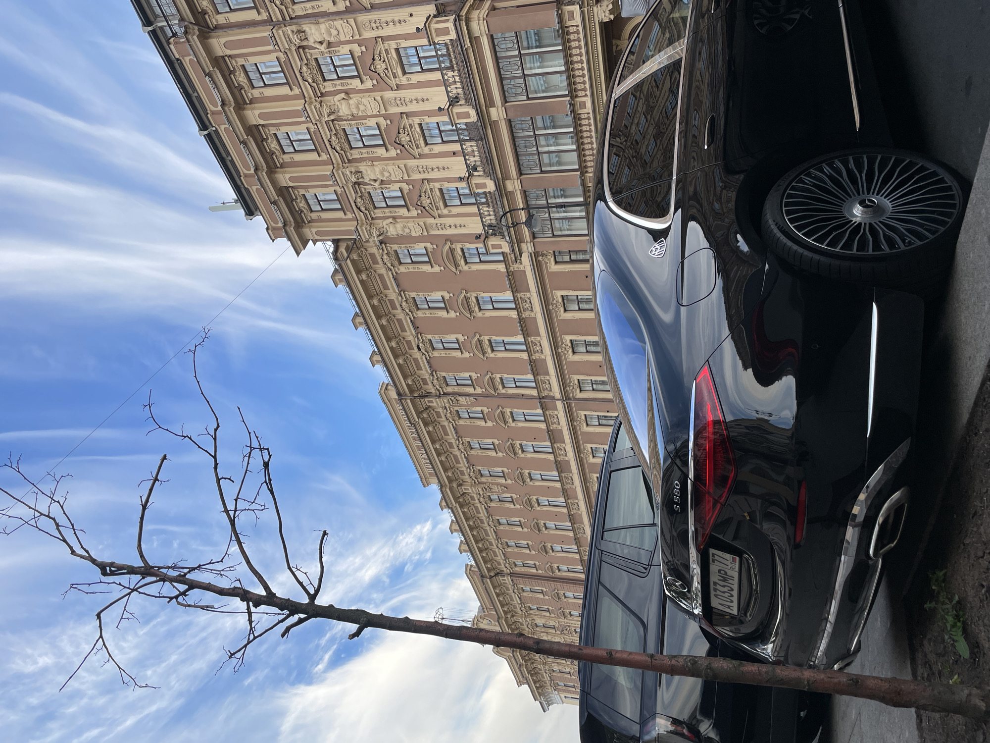 An outdoor, daylight scene rotated 90 degrees counter-clockwise captures a street view from a low angle, featuring an ornate building, a parked luxury car, and a bare tree branch against a bright sky.
In the upper right, a multi-story, classical-style building with a light brown facade is prominent. It features numerous windows, elaborate decorative stucco work, pilasters, and balconies. Reflections of the building are visible on the surfaces of the parked car. Partially visible text on the building reads "HOTEL d'EUROPE".
A dark-colored (likely black or dark blue) Mercedes-Benz S-Class sedan, specifically an S 580 as indicated by the emblem on the trunk, is parked along the curb. A Maybach emblem is visible on its side. The car's polished surface reflects the sky and the intricate building. It has distinctive multi-spoke alloy wheels. The rear license plate, "A 033 MP 77 RUS", is clearly visible, indicating it's a Russian vehicle, with "77" being a Moscow region code.
A leafless, bare tree branch extends across the middle left of the frame, silhouetted against the sky. The sky itself is a clear blue with delicate, wispy white clouds stretching horizontally.
The image depicts a quiet urban scene, likely taken during the day given the bright, natural light. The architectural style, the visible "HOTEL d'EUROPE" sign, and the Russian license plate align with the provided context of Saint Petersburg, Russia.