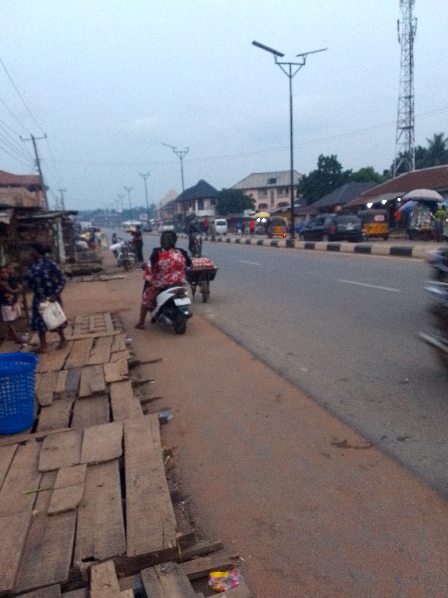 This image captures a bustling street scene in Orlu, Nigeria, under an overcast sky, suggesting late afternoon or dusk. The setting is an urban or semi-urban commercial area, with a paved road, roadside stalls, and various buildings.
On the left, a makeshift wooden platform serves as a walkway or market space, with a bright blue plastic basket in the foreground. Two women and a child are visible near these wooden structures; one woman in a blue and white patterned dress carries a white bag.
In the mid-ground, a woman in a red and white patterned outfit and a dark head covering sits on a white scooter or moped, facing right. Next to her, a person pushes a handcart or wheelbarrow filled with what appears to be red produce. The road features a solid white line and black and white striped kerbs. Several vehicles, including cars and yellow auto-rickshaws (locally known as Keke Napep), are parked or moving along the street. A blurred object on the far right indicates a fast-moving vehicle.
Modern streetlights with flat, wide fixtures are spaced along the road, and power lines are visible overhead on the left. In the background, a mix of multi-story and single-story buildings with various roof types can be seen. A prominent cellular or telecommunications tower stands tall on the right side of the horizon. The overall scene depicts typical daily life and commerce in a busy Nigerian town. No legible text is visible in the image.