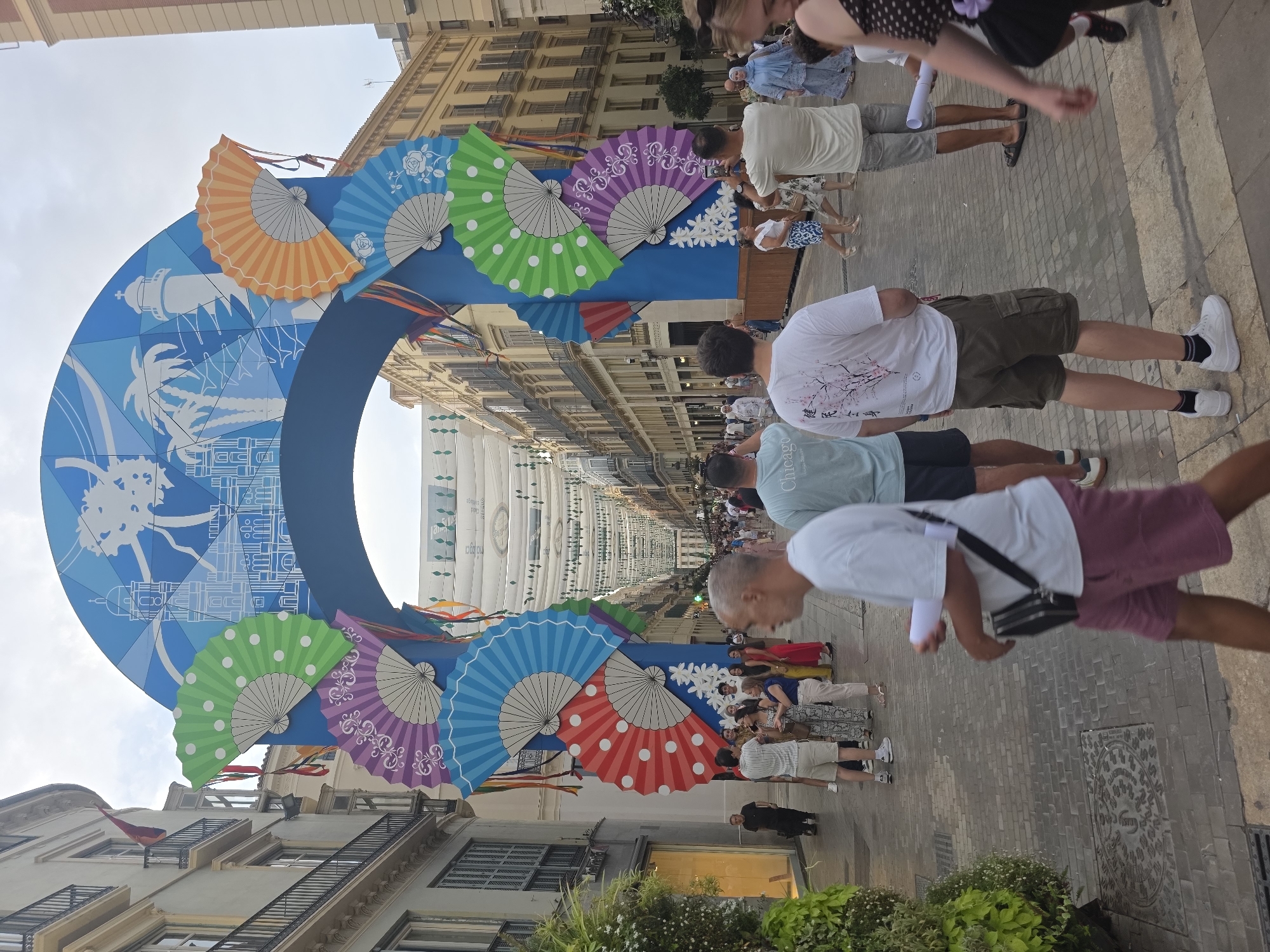 The image depicts a festive street scene in Malaga, Spain, likely during a local celebration or fair. A large, vibrantly colored archway, decorated with stylized imagery of palm trees and buildings, spans the width of the street. Adorning the arch are numerous large, decorative fans in various colors and patterns, including polka dots and floral motifs. The street is paved with cobblestones and lined with multi-story buildings, typical of Spanish urban architecture, with balconies and shops visible.
A crowd of people are walking along the street, some looking at the decorations, others engaged in conversation. Several individuals are captured in motion, suggesting a bustling atmosphere. Among the visible people are men and women dressed in casual summer attire. One man in the foreground is wearing a white t-shirt with Japanese lettering and dark cargo shorts, holding a white rolled-up paper. Another man to his right wears a light blue t-shirt and grey shorts, also holding a rolled-up paper. Further down the street, more people are milling about, with some taking pictures.
Overhead, string lights and decorative elements hang across the street, enhancing the festive ambiance. The sky is overcast, indicating a cloudy day. The overall impression is one of a lively public space filled with people enjoying an event. The distinctive architecture and the style of the decorations strongly suggest Malaga, a city known for its vibrant street life and cultural events.