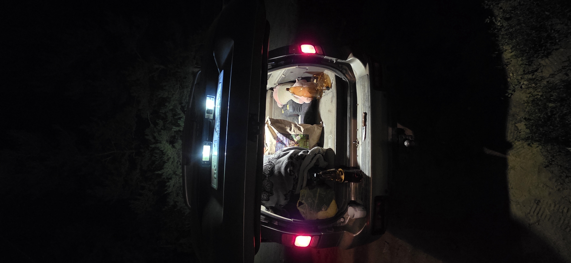 The image shows the open trunk of a car at night. The car is parked in an outdoor, unlit area, possibly a rural or undeveloped location, with trees visible in the background. The trunk contains several items, including bags of groceries, a blanket, plastic bottles, and a knife. The car's brake lights are illuminated, casting a red glow. The scene appears to be in the city of Karakol, Kyrgyzstan, though no specific landmarks or identifying features of the city are visible.