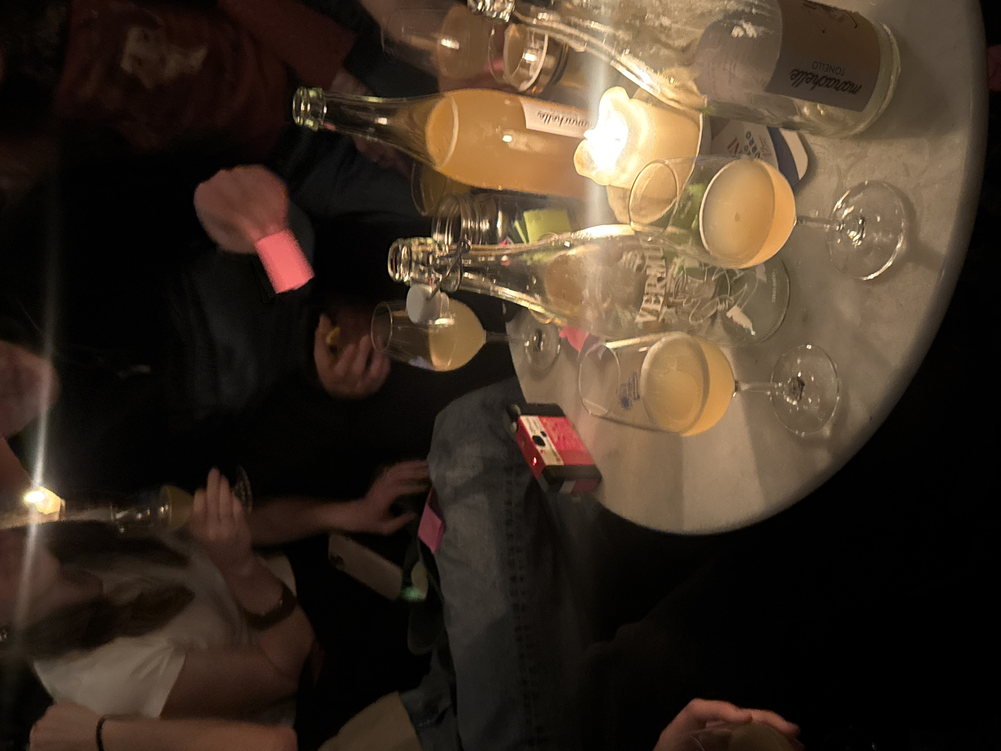 The image shows a group of people gathered around a small, round, light-colored table in a dimly lit setting, likely a bar or restaurant. On the table are several wine glasses filled with an amber-colored liquid, possibly cocktails. There are also several empty wine bottles, including one labeled "mariachelle TONELLO". A lit candle sits among the glasses and bottles, providing additional light. People are holding and drinking from the glasses, and their hands and arms are visible around the table. One person is wearing a bright pink wristband. A red object and a smartphone are resting on one person's lap. The environment suggests an intimate, social gathering. It's not possible to ascertain the location in Amsterdam, Netherlands.