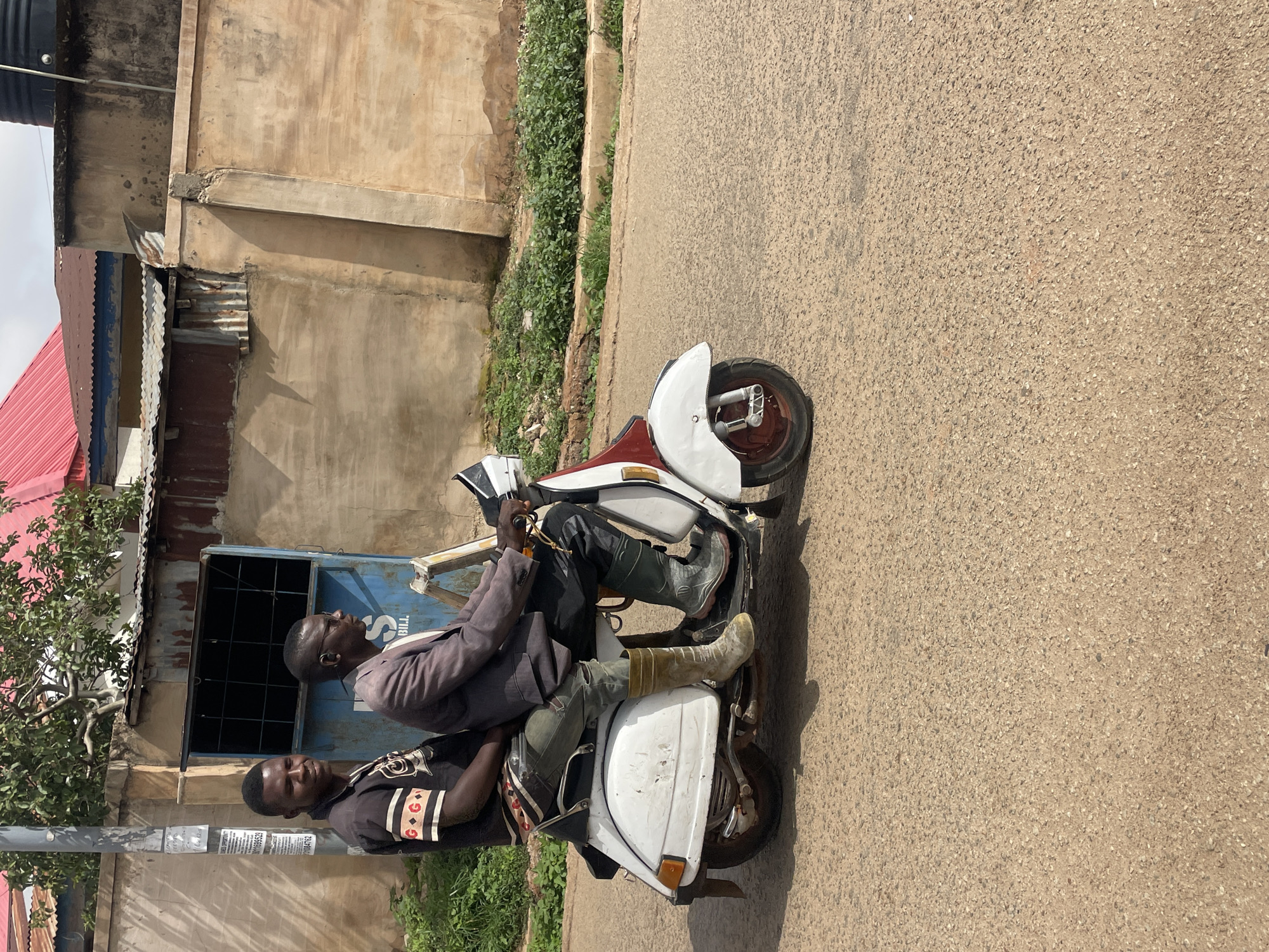 Two African men are on a white and burgundy vintage Vespa scooter parked on a paved street. The driver wears a gray jacket, black pants, dark-colored boots, and glasses. The passenger wears a black shirt with decorative stripes on the sleeves and black pants, and he is behind the driver. The backdrop includes an old building with corrugated metal sheets, a blue door, and some vegetation. The location is Jos, Nigeria.