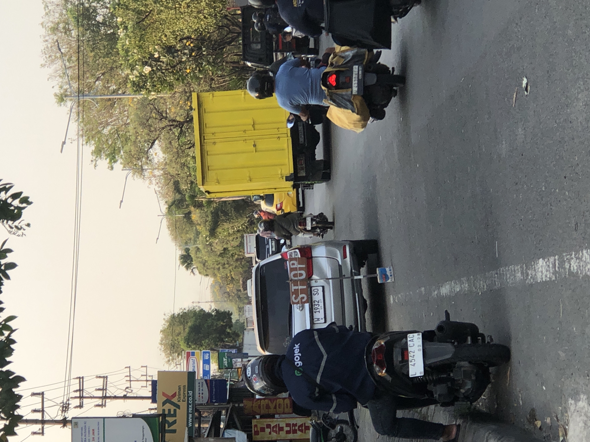 In Ngaban, Indonesia, this scene captures a street view with various vehicles and pedestrians under a bright, overcast sky. In the foreground, a person on a black motorcycle, wearing a dark blue jacket with the "gojek" logo, is stopped at the curb. The motorcycle has a license plate that reads "L 4542 CAC".

Further ahead in the street, a silver SUV is stopped. A "STOP" sign is attached to its rear, propped up by a blue and white container on the asphalt. The SUV's license plate reads "W 1932 SO". Behind the SUV, a yellow box truck is visible, and a bright yellow car is in the lane next to it. A person on a motorcycle is seen riding behind the yellow box truck.

Other individuals on motorcycles are also present, some with deliveries or luggage. The environment includes power lines overhead, trees lining the street, and various signs for businesses along the roadside, including "REX Kiriman Express". The road surface is asphalt, marked with a white line. The time of day appears to be late afternoon due to the warm light.