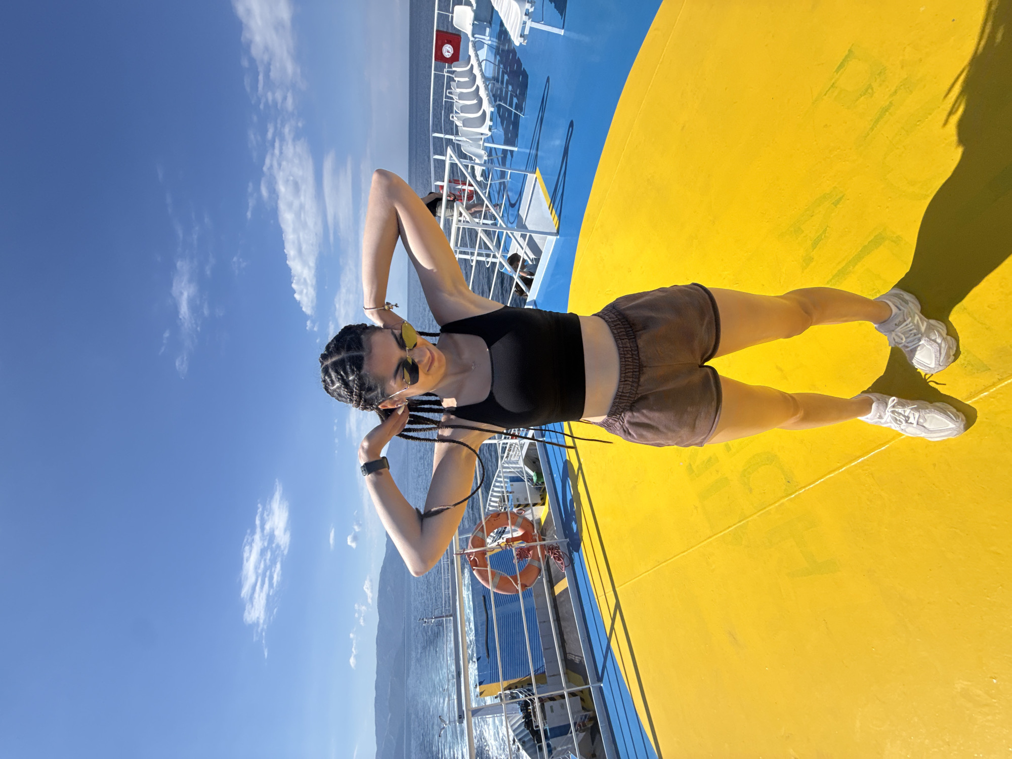 A woman with dark braided hair stands on the deck of a ferry with her arms behind her head. She is wearing sunglasses, a black crop top, brown shorts, and white sneakers. The deck is painted bright yellow and blue. In the background, there is a railing, some white chairs, and the sea under a clear blue sky with scattered clouds. A life preserver is visible on the railing. The woman's shadow is cast on the yellow deck. The location is the sea, near the coast of Greece.