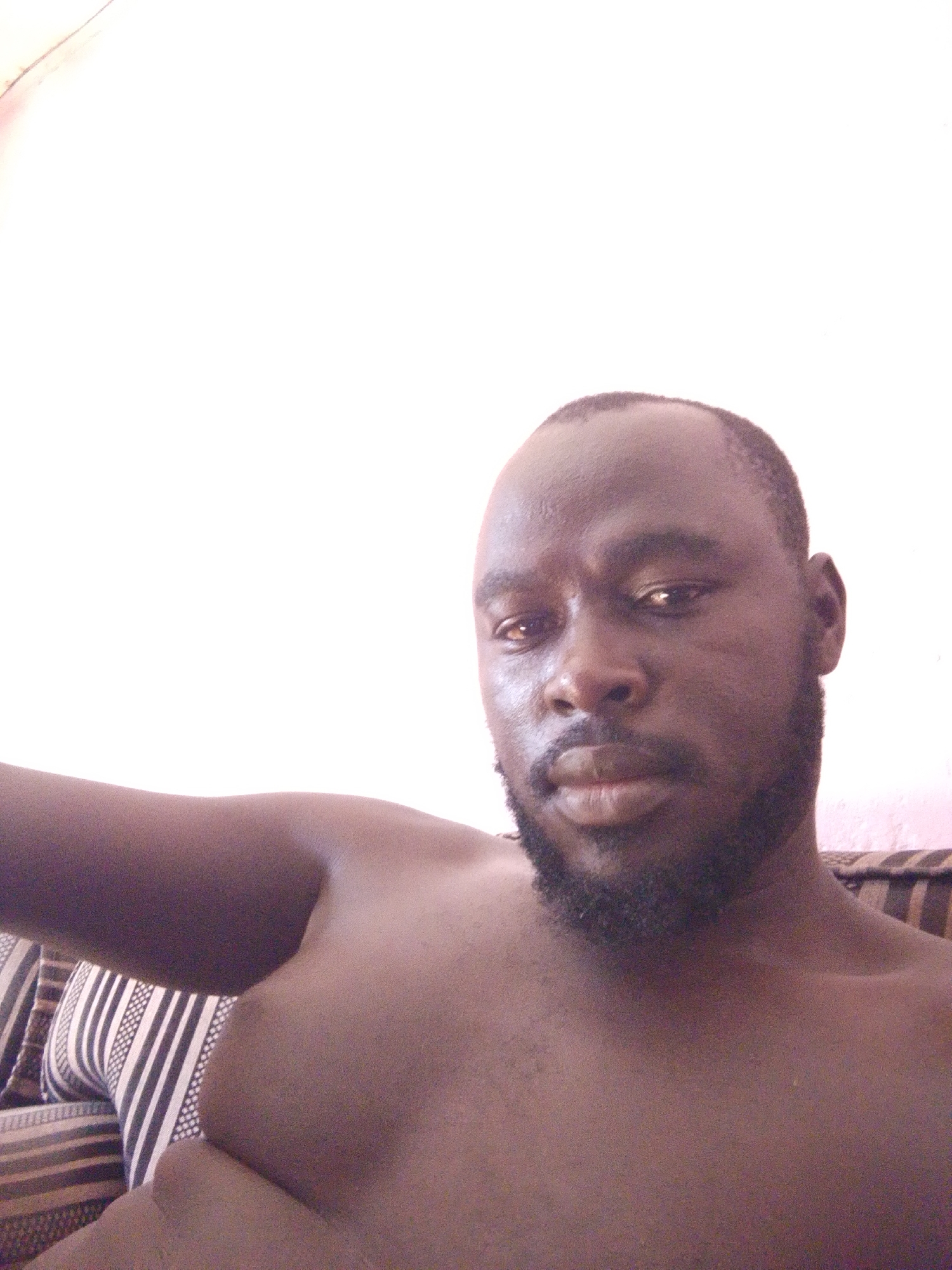 A medium-close-up shot presents a shirtless Black man with a dark beard and short hair. He is positioned in front of a light-colored wall and appears to be reclining on a striped cushion or sofa. There are no other visible people, objects, or actions. The environment is limited to the immediate background and furniture. The location is not discernable from the image.